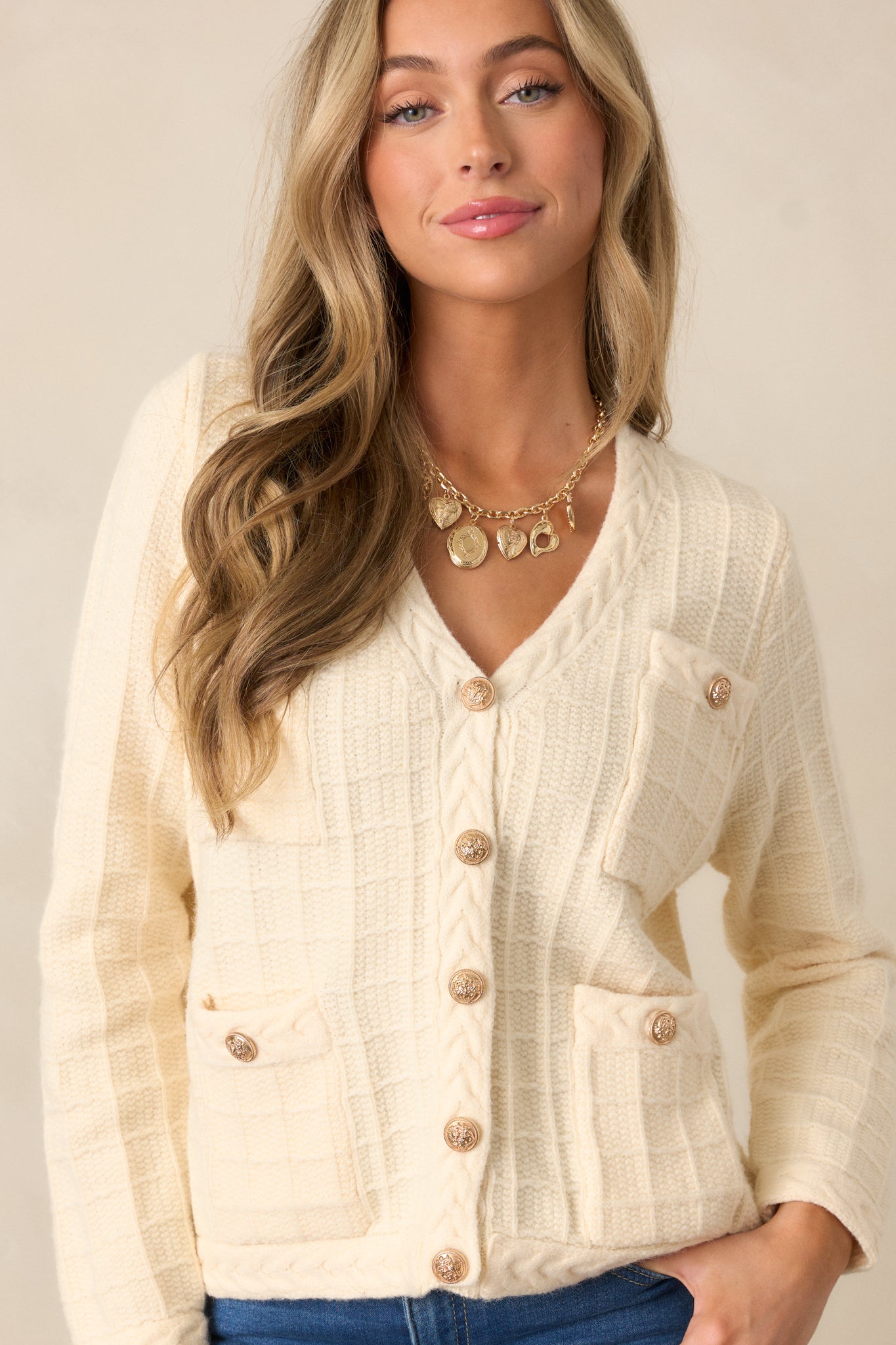 A close-up of the top half of the ivory cardigan, emphasizing the v-neckline, cable knit grid pattern, and the functional gold buttons along the front.