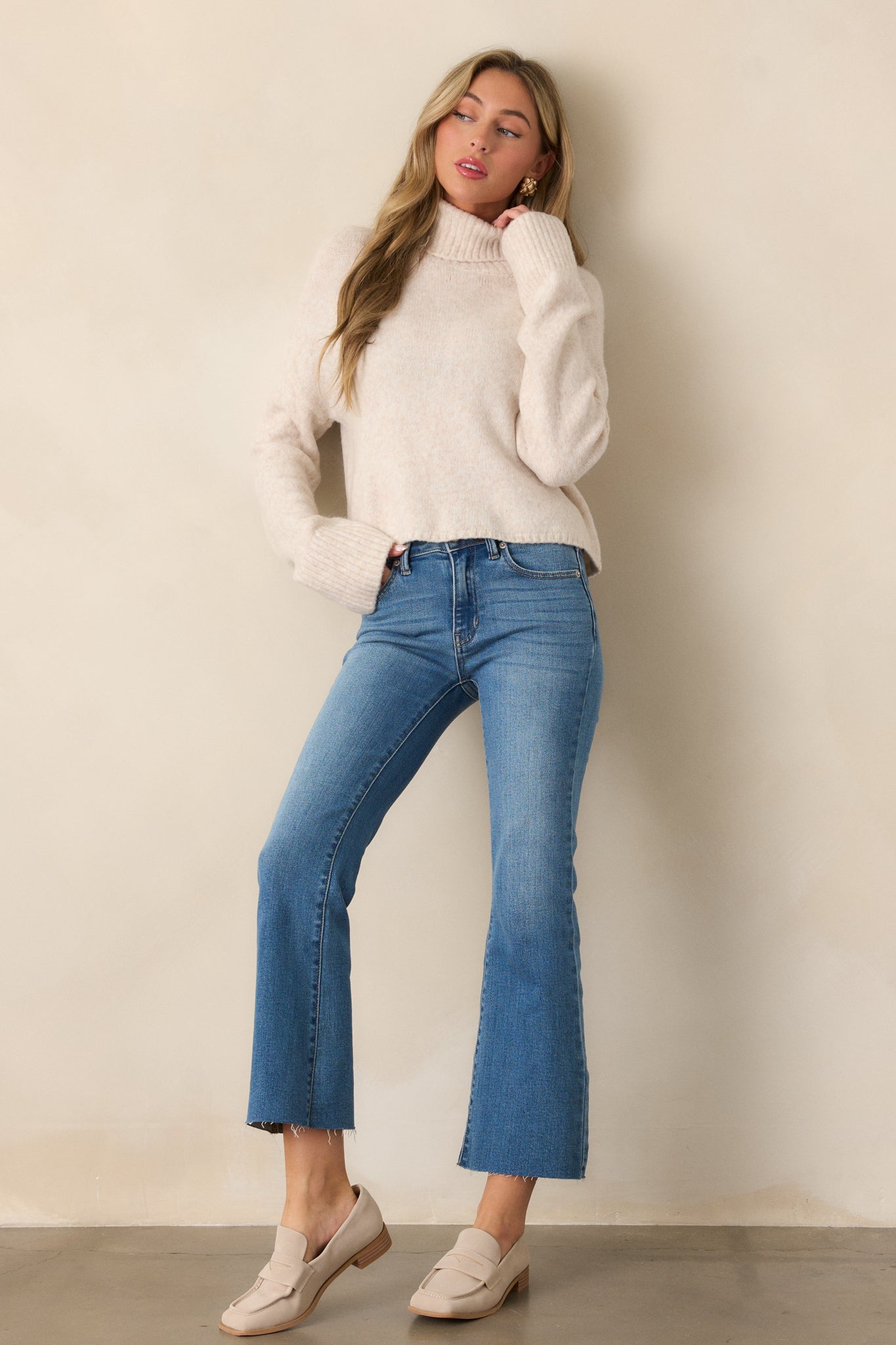 Slightly angled view of a light oatmeal sweater showing its relaxed fit, ribbed turtleneck, and long sleeves with thick ribbed cuffs and bottom hem detailing.