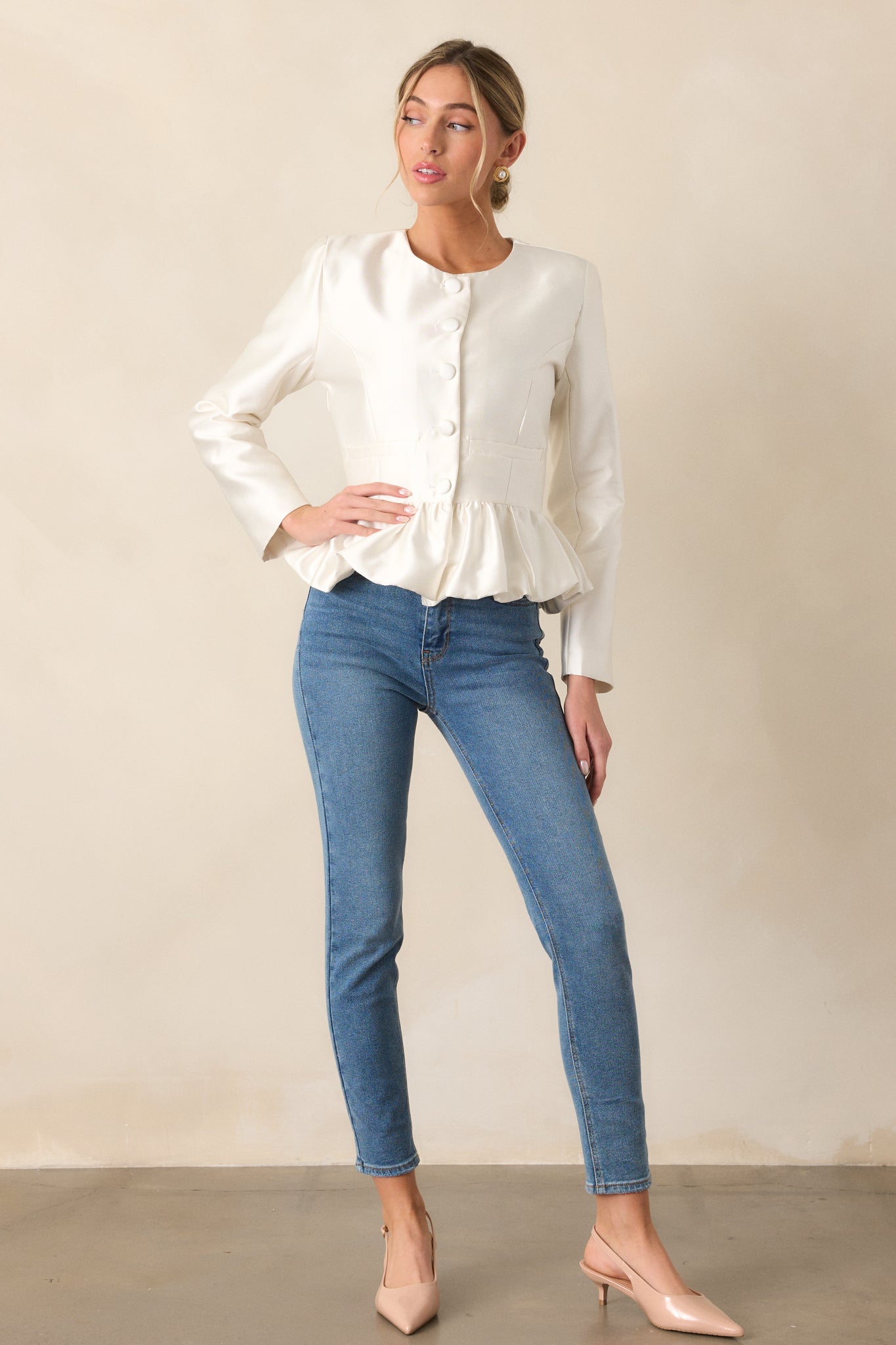 A full-length view of the ivory top highlighting the peplum silhouette, long sleeves, and thick satin fabric, with a focus on the button front and faux waist pockets.