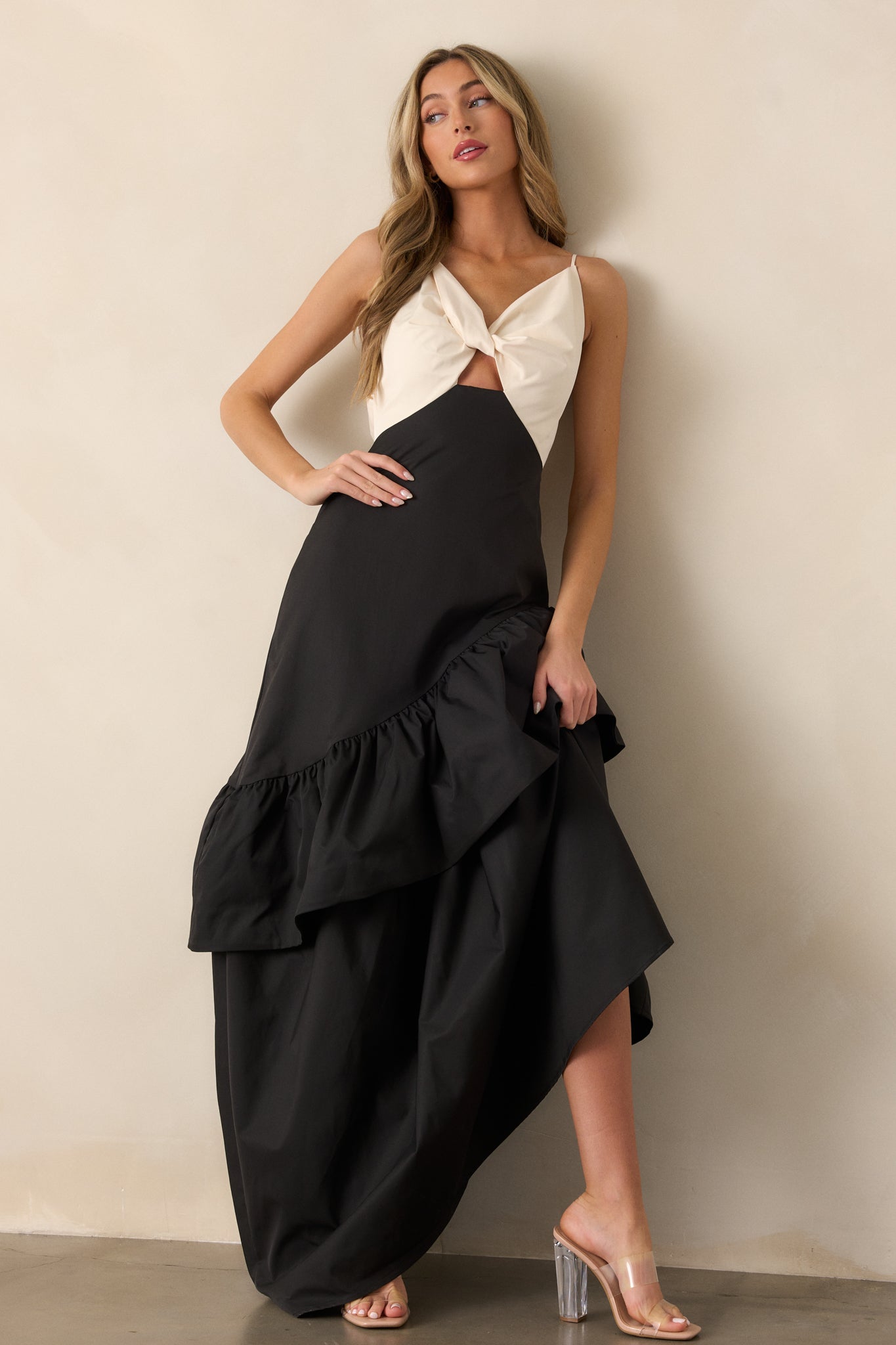 Full view of a black dress featuring an ivory v-neck top with twist detailing, a drop waist design, and an asymmetrical hem with ruffled accents.