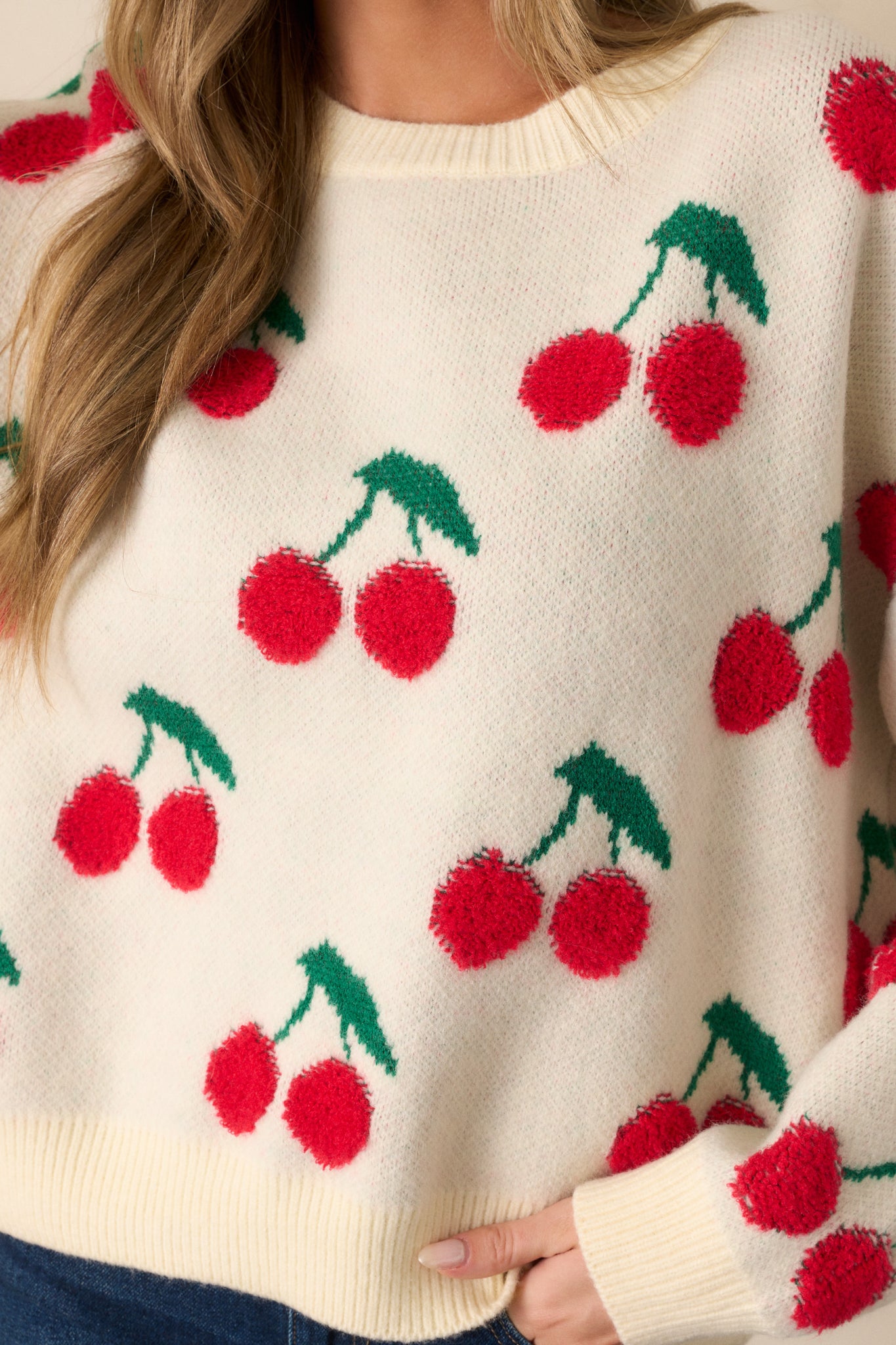 A detailed close-up showing the cherry embroidery on the ivory sweater. The ribbed texture of the crew neckline and sleeve cuffs is visible, adding depth and dimension to the relaxed fit.