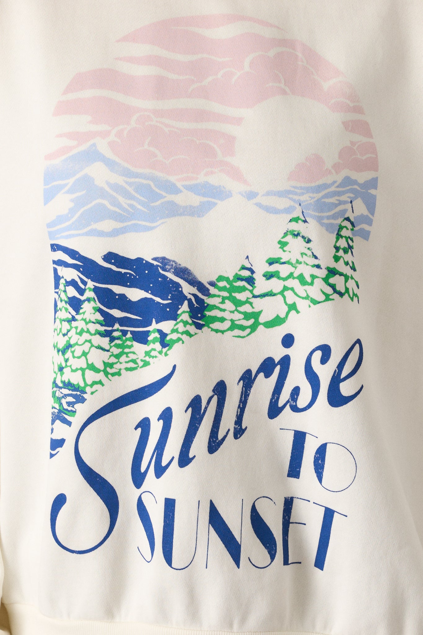 A close-up of the ivory sweatshirt’s 'Sunrise to Sunset' snow-covered mountain landscape graphic. The soft, 100% cotton brushed fleece fabric and the crew neckline are clearly visible, showcasing the top's comfortable and stylish look.