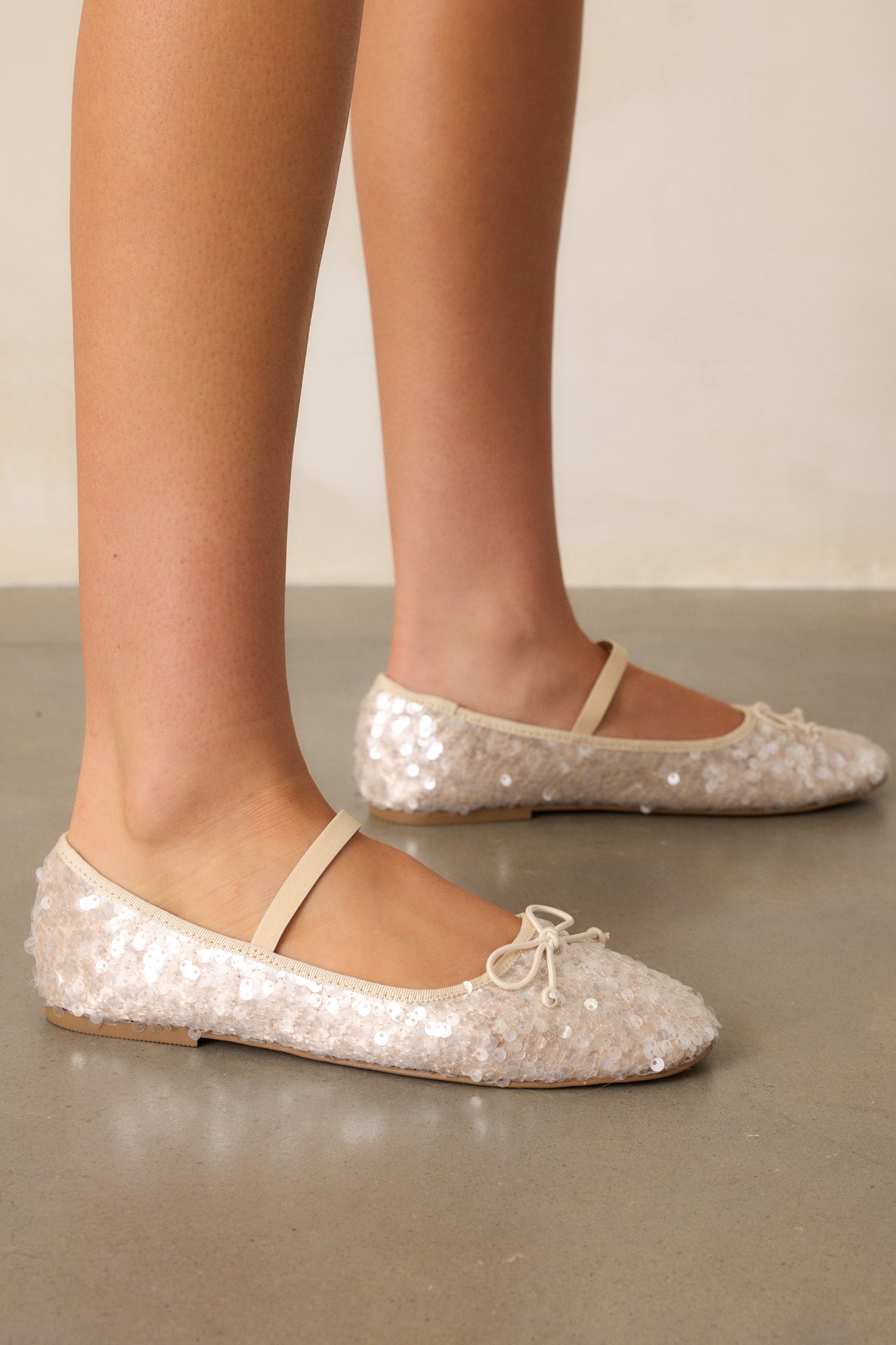 Beige sequin flats with a rounded toe, highlighted with a small beige bow and an across-the-foot strap, shown from an angled side view.