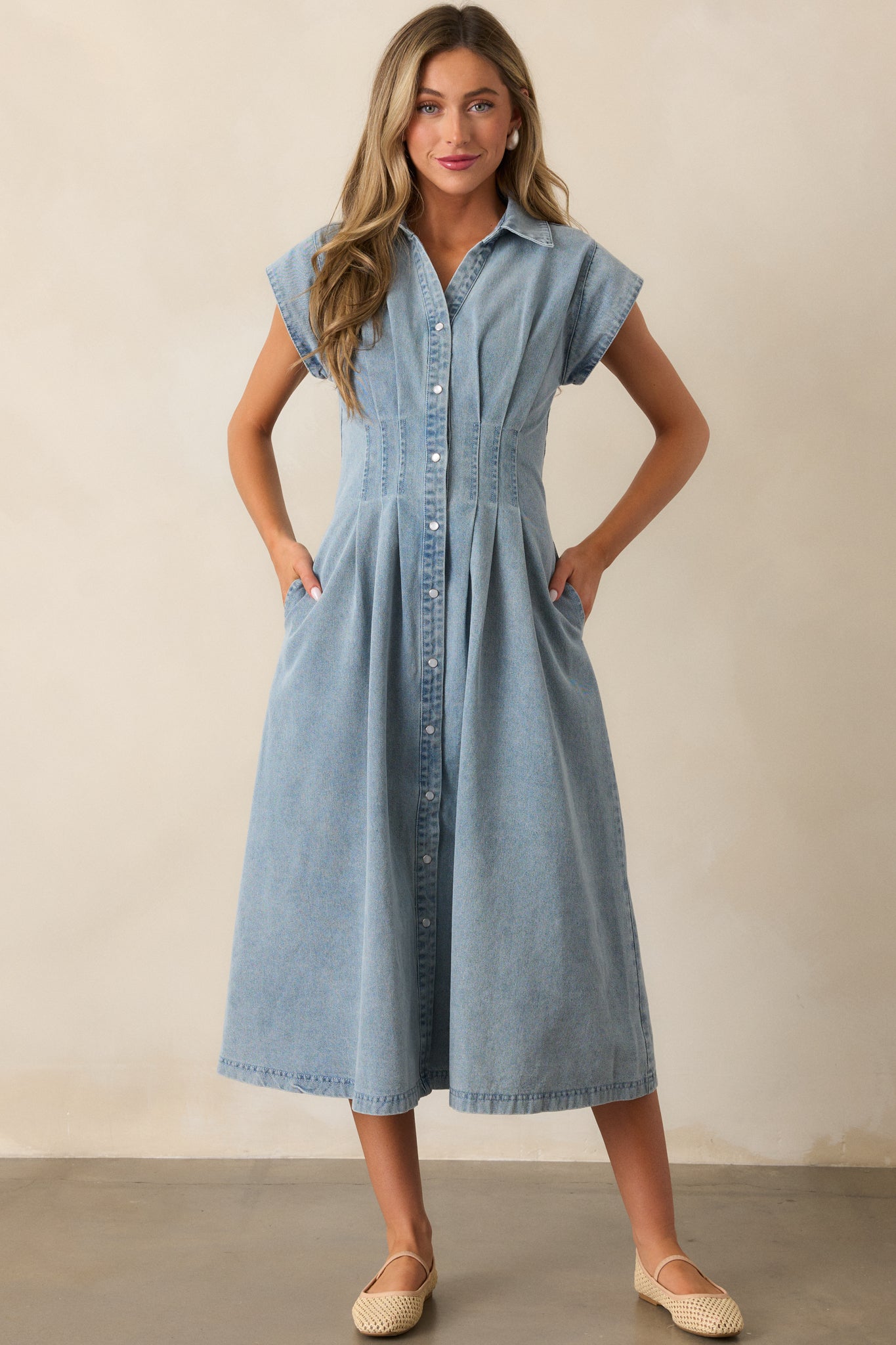 Light wash denim dress with cap sleeves, pearlized snap button front, and pleated waist detail, styled with two functional pockets.