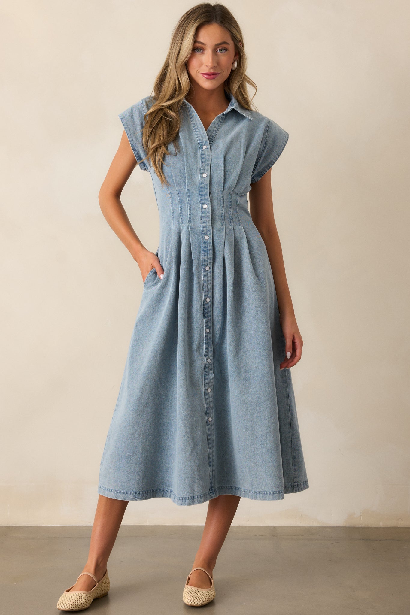 Denim dress in a light wash featuring a button-up front with pearlized snaps, cap sleeves, and a pleated design around the waist.