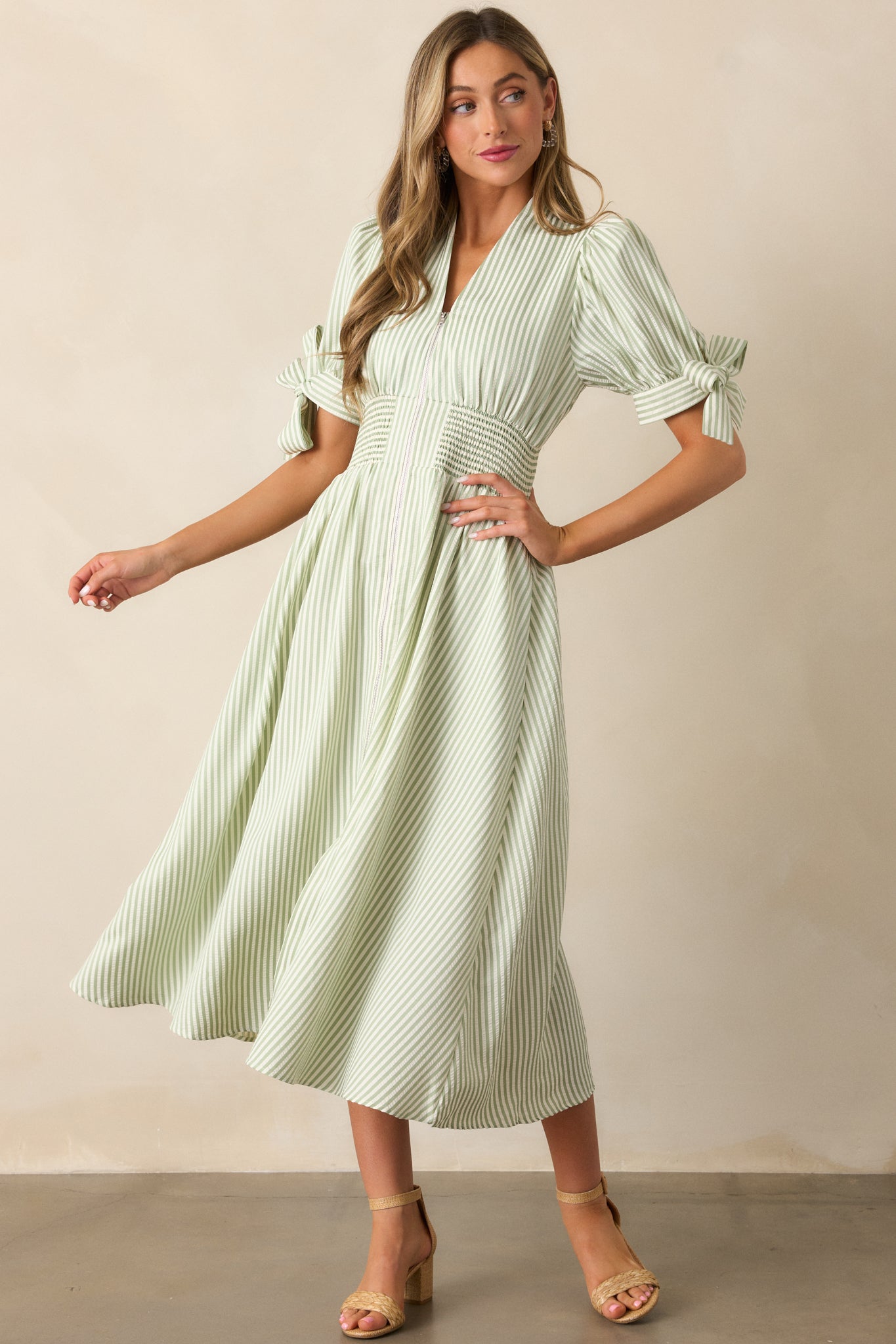 Green stripe dress with a smocked waist, functional pockets, and a V-neck neckline that transitions into a front zipper, featuring tie details at the sleeve ends.