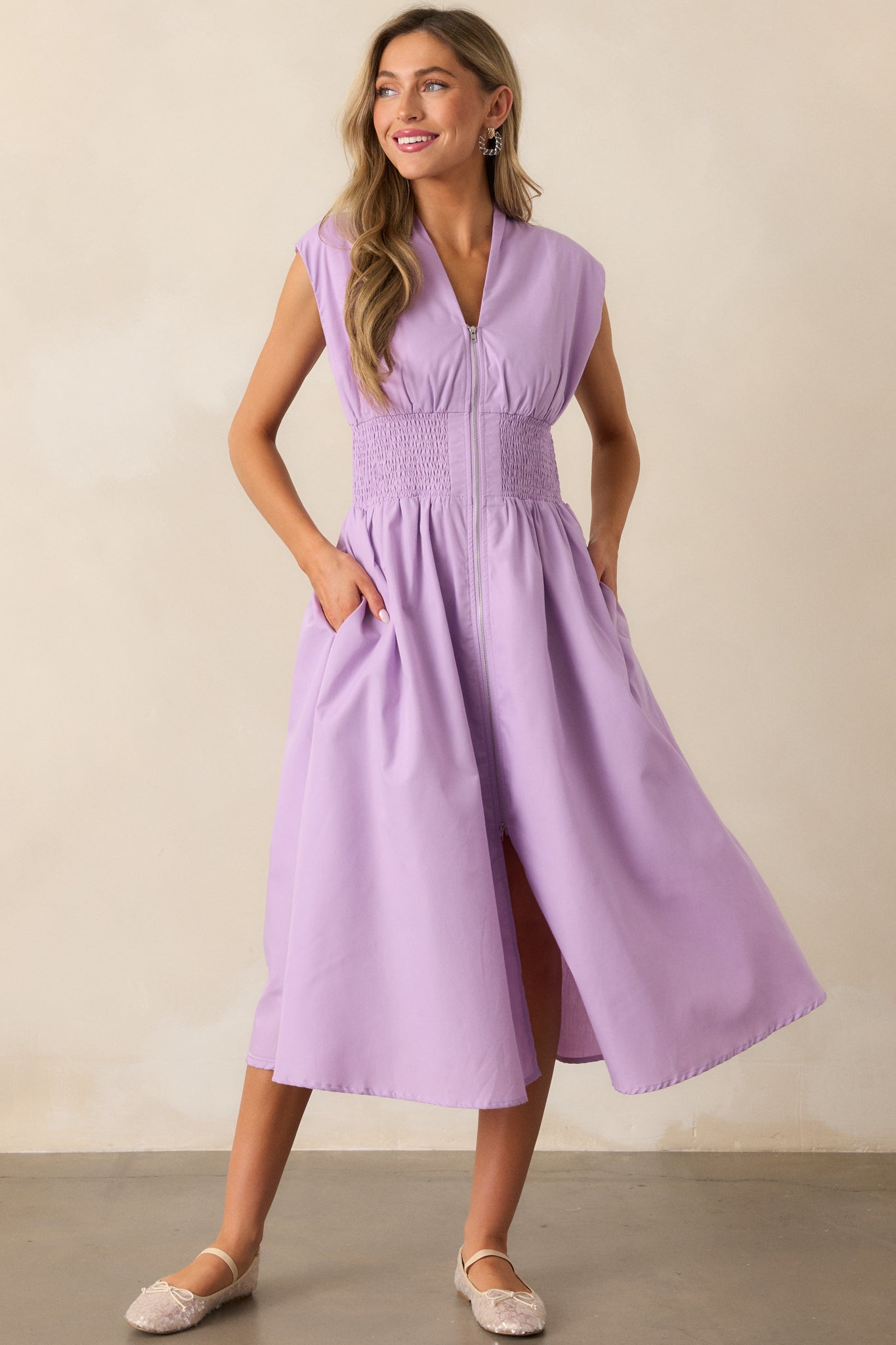 This lavender midi dress features a chic v-neckline, capped sleeves, a front slit, and functional hip pockets, all highlighted by its comfortable smocked waist.