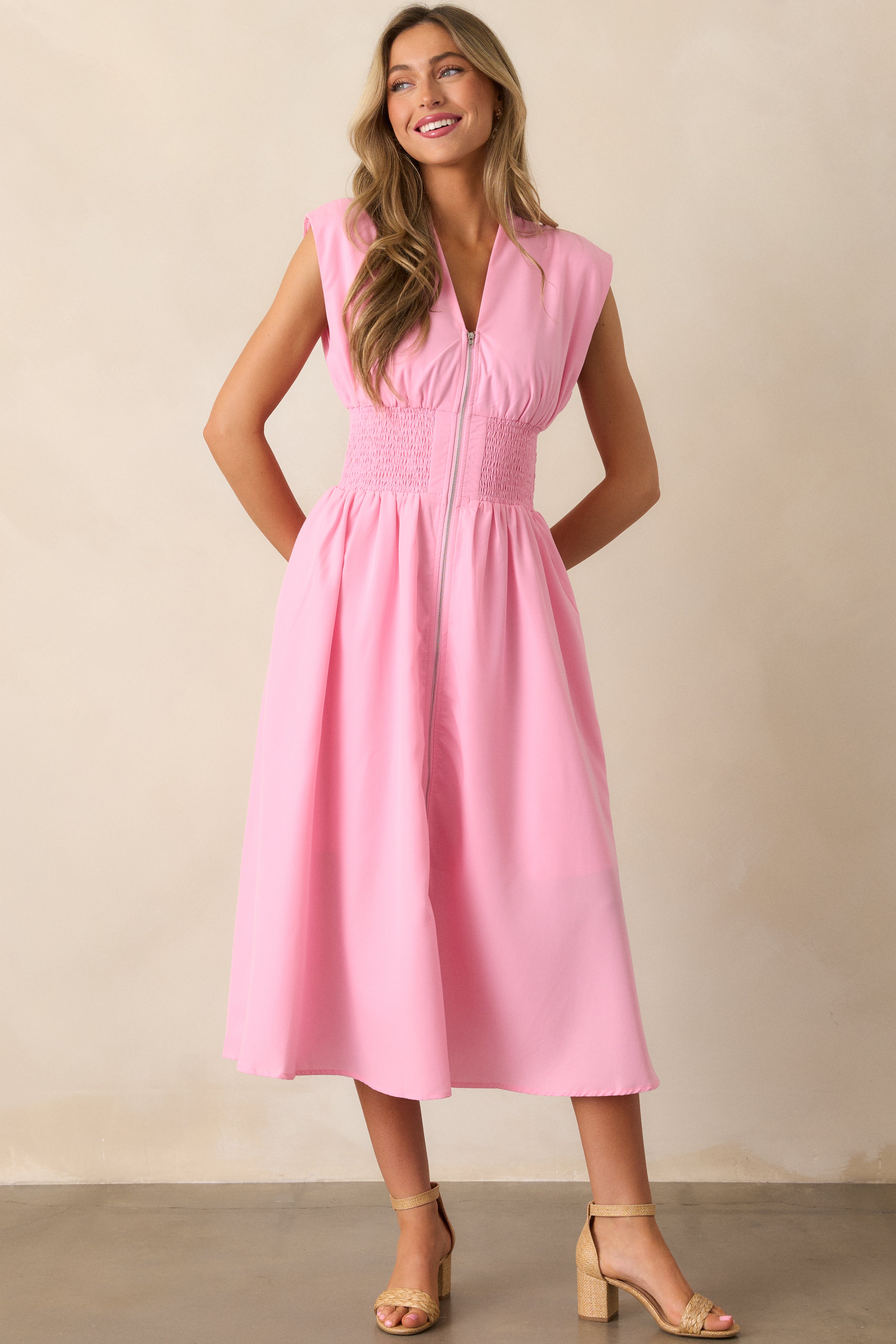 Front angled view of a baby pink midi dress featuring a v-neckline, padded shoulders, a full zipper front, a fully smocked waist, functional hip pockets, and a front slit.