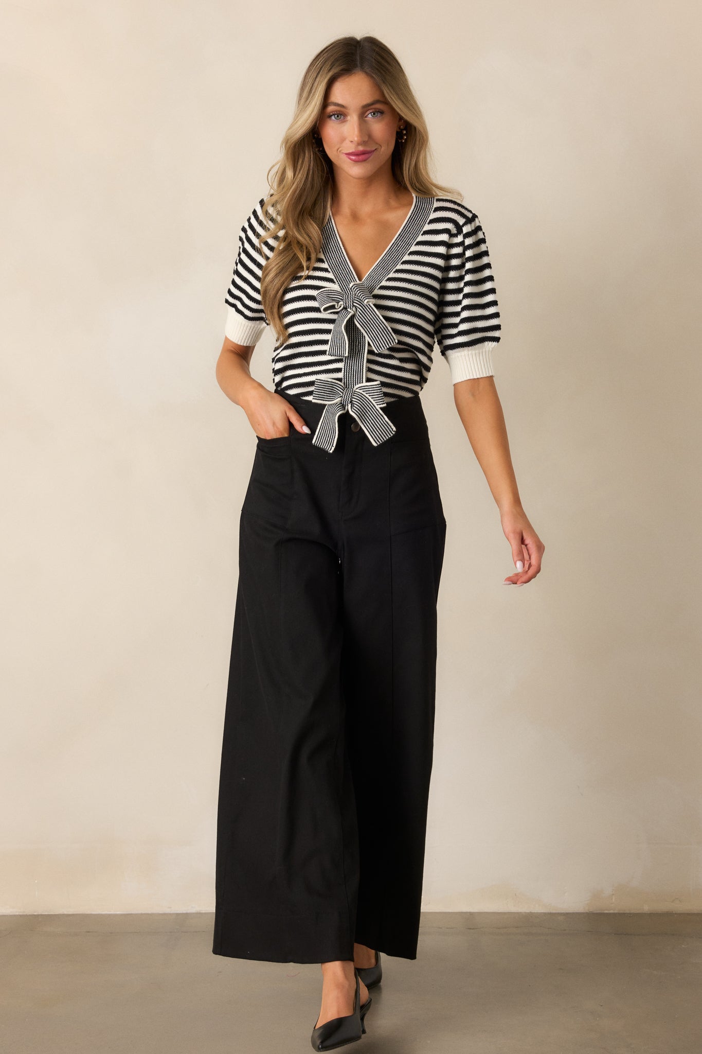 A black top with a v-neckline and black and white stripe print, featuring puff sleeves with ribbed cuffs and a bow front detail, shown in a full view.