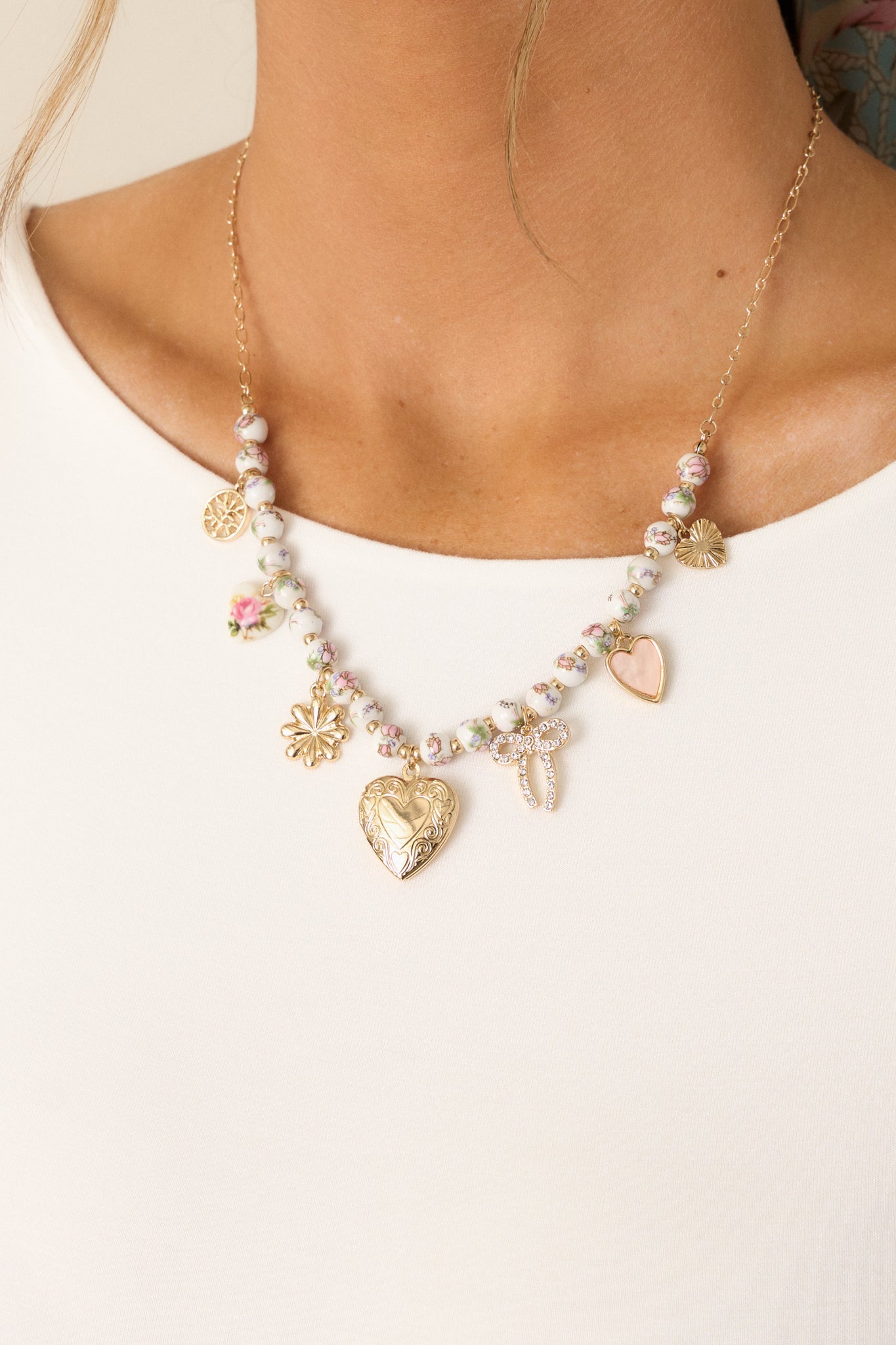 A close-up of the gold and ivory charm necklace, highlighting the thin gold chain with ivory and pink floral beading, heart charms, and a heart locket hanging at the center.