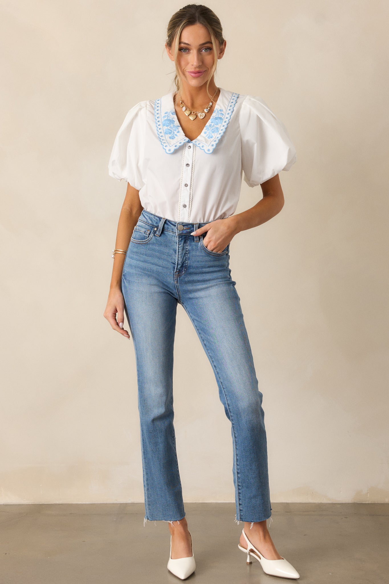 A white top with a peter pan collared neckline featuring blue floral embroidery, a button front, and short puff sleeves.