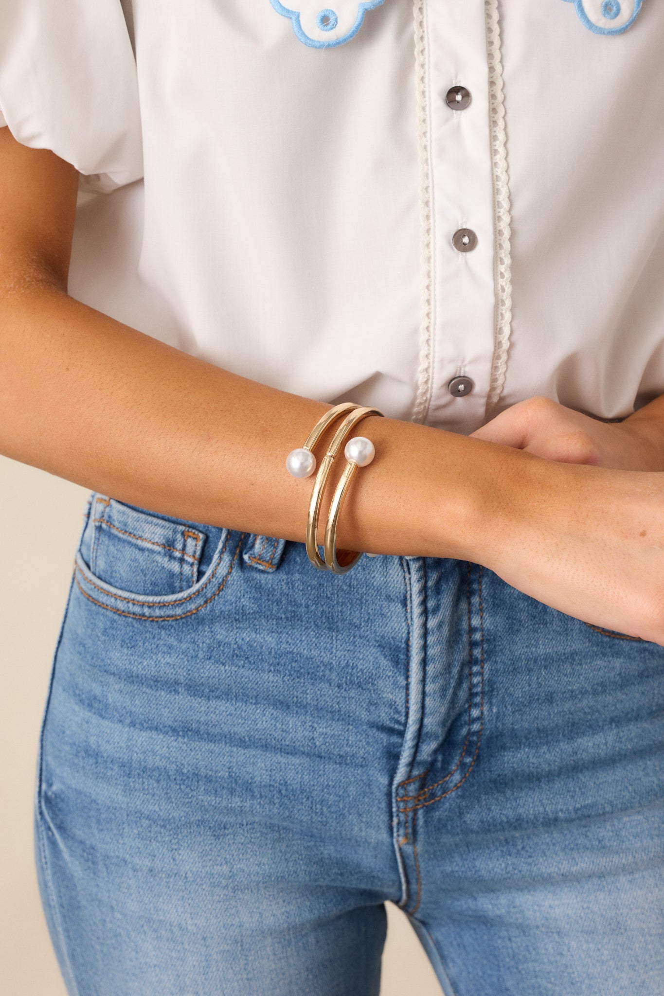 A cropped view of the gold bracelet, showing its thick gold hardware, layered bangle design, and one large ivory faux pearl at the end, with the snap closure partially visible.