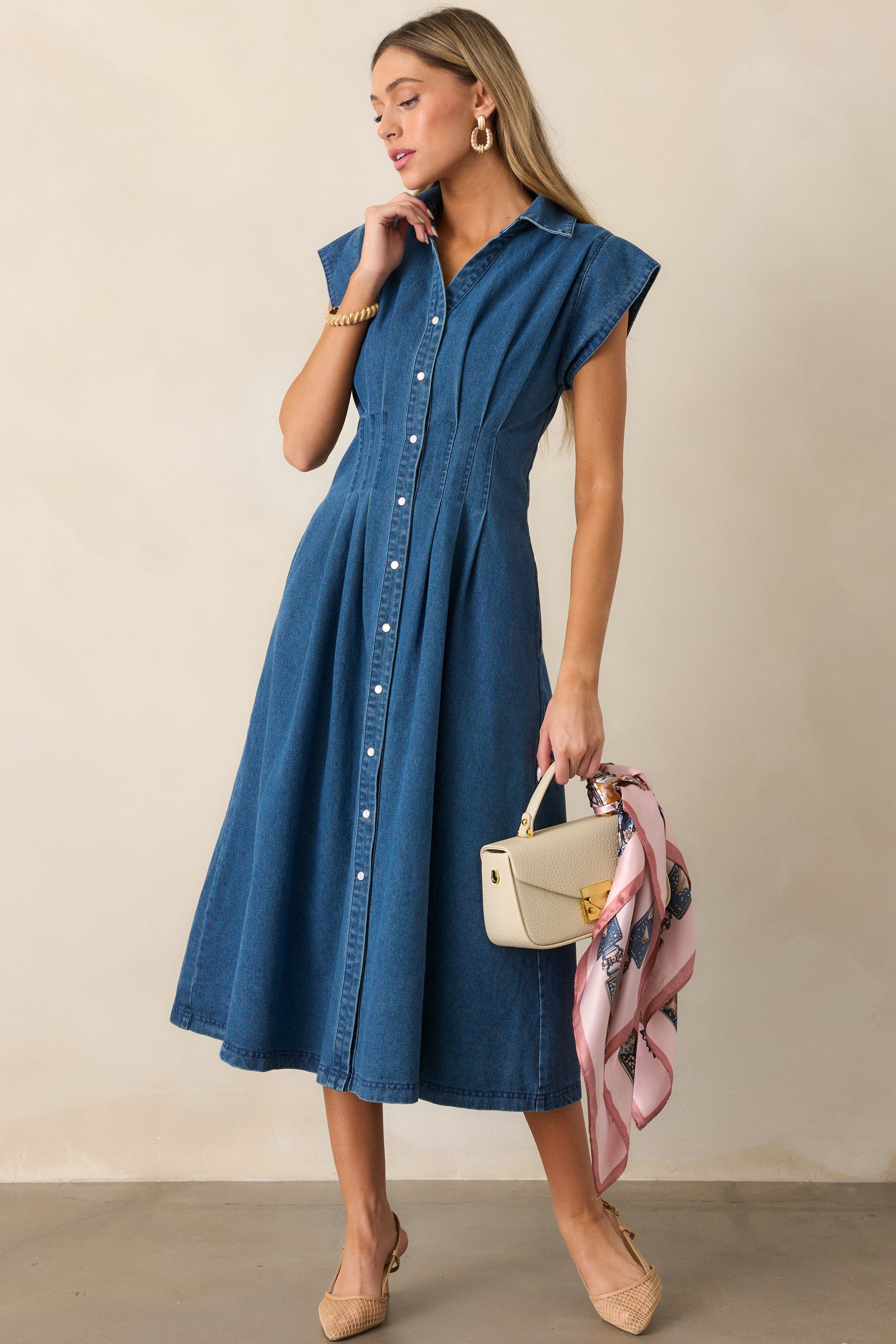 Dark wash denim dress with cap sleeves, pearlized snap button front, and pleated waist detail, styled with two functional pockets.