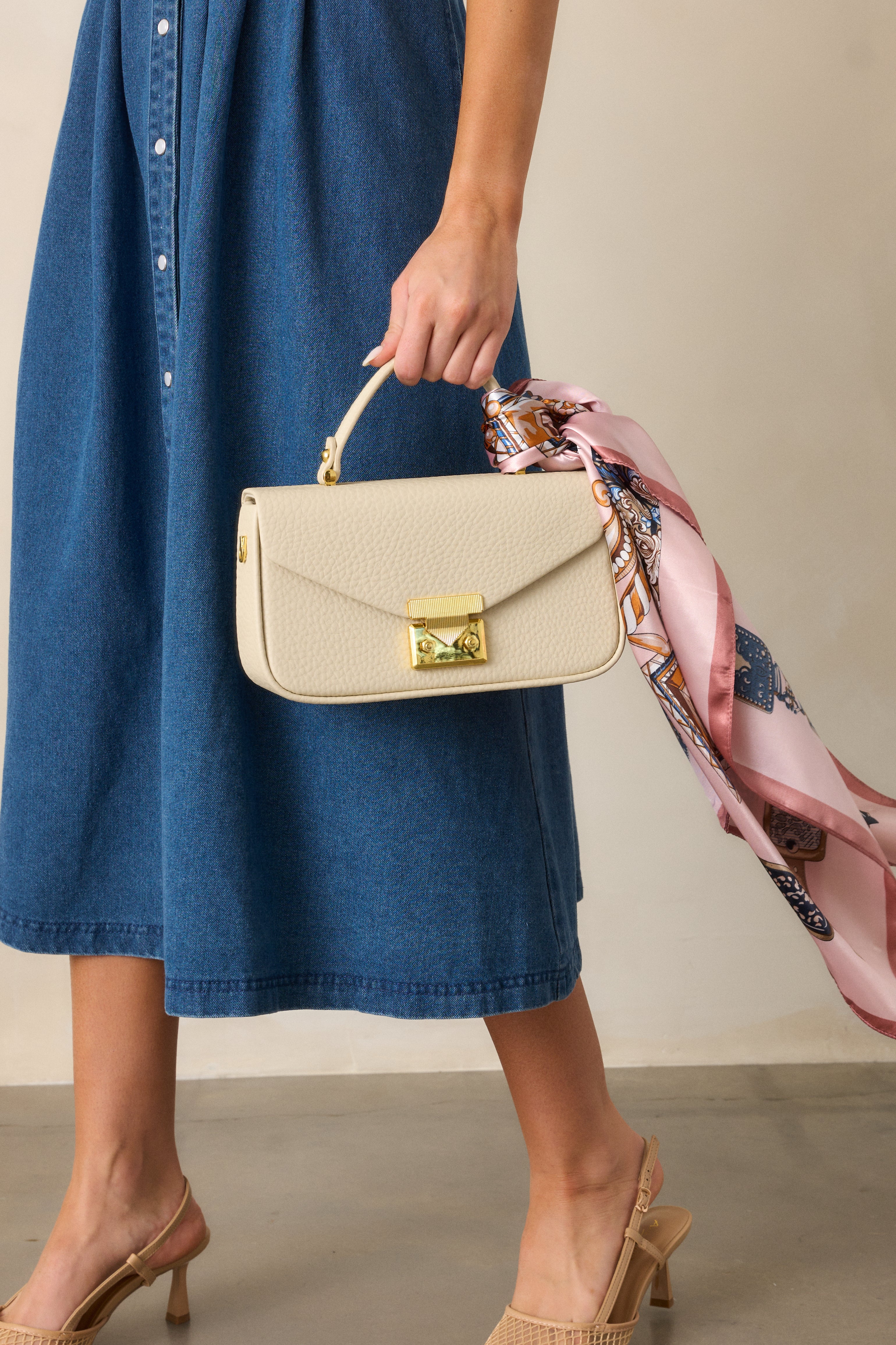 Full view of an ivory handbag with a rectangular shape and gold hardware, featuring a gold triangular clasp tuck lock, carried by the handle.