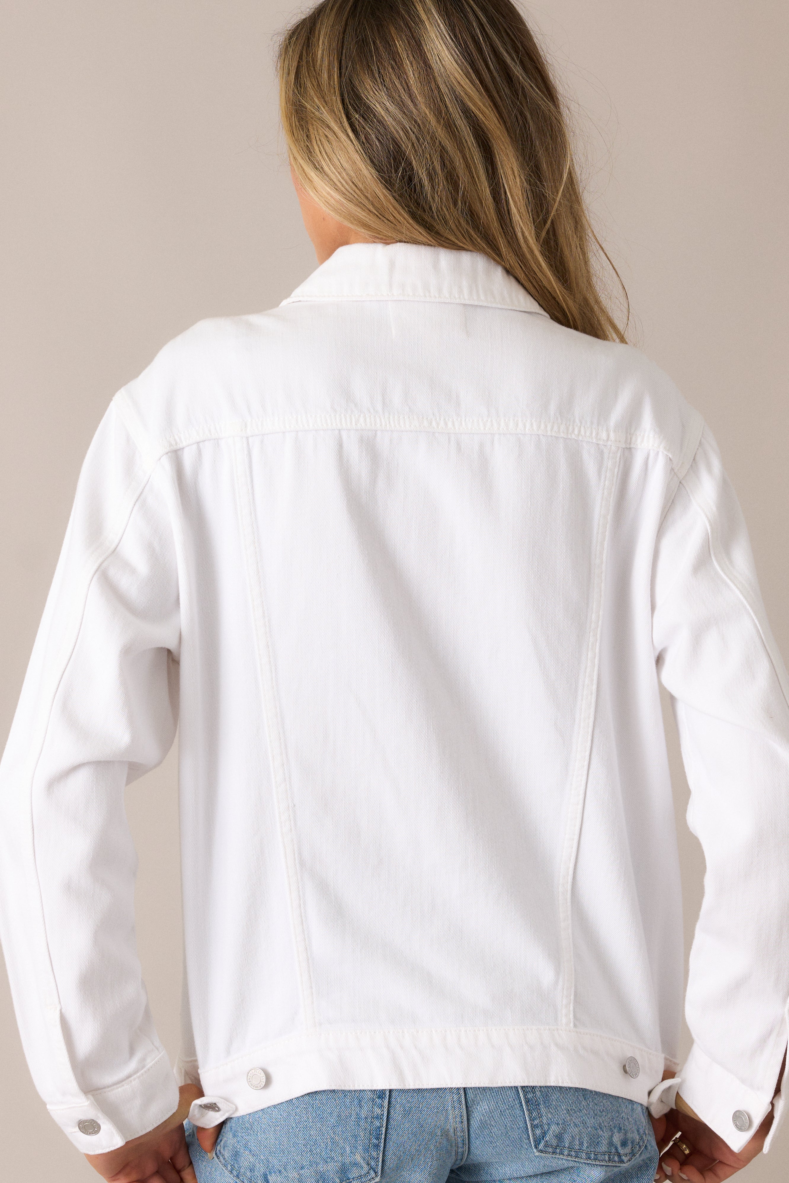 The white denim jacket from the back, showcasing the clean lines and buttoned sleeves that give the jacket a tailored, yet casual feel.