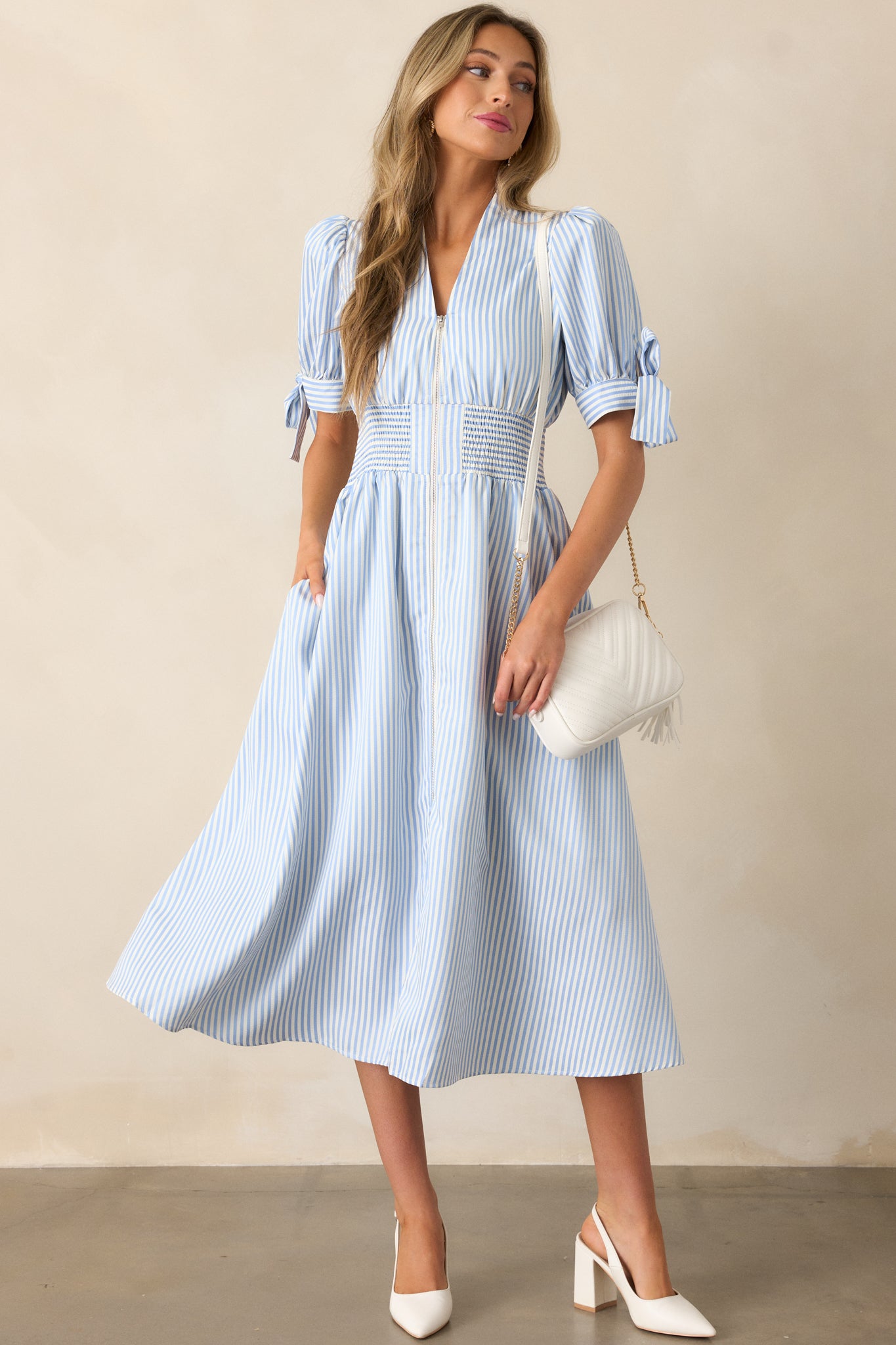  Blue stripe dress with a smocked waist, functional pockets, and a V-neck neckline that transitions into a front zipper, featuring tie details at the sleeve ends.