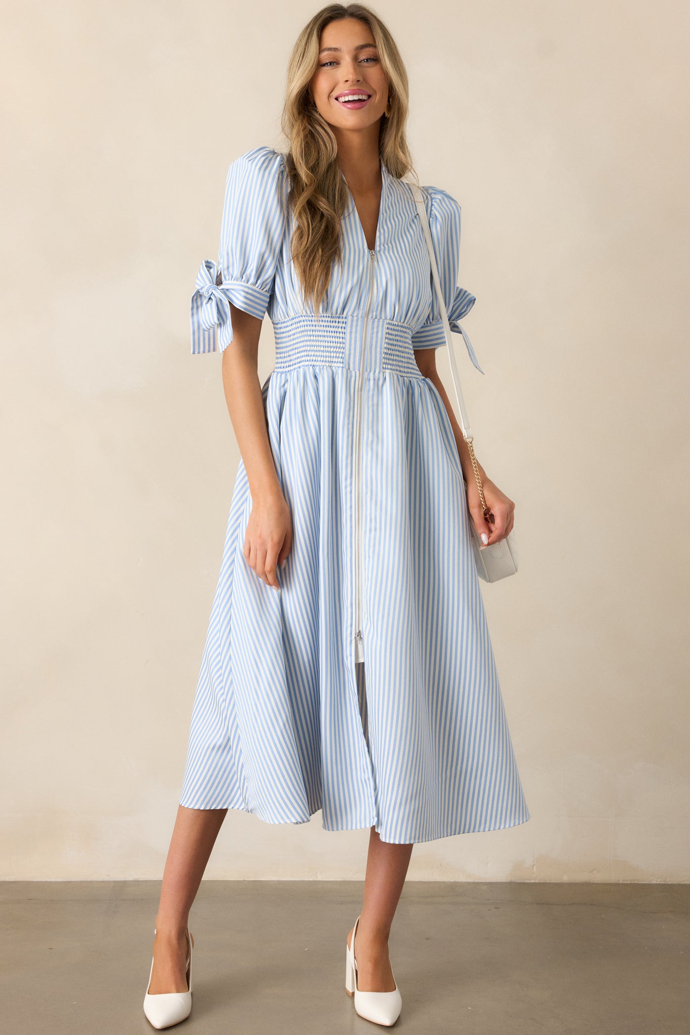 Dress in a blue stripe pattern, highlighting the fitted smocked waist, front zipper, and sleeve ties, along with two functional pockets.