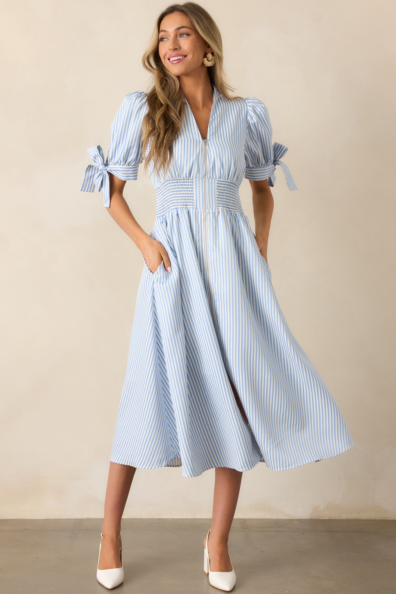 Blue stripe dress with a smocked waist, functional pockets, a V-neck neckline, and tie details on the long sleeves, styled with a front zipper.