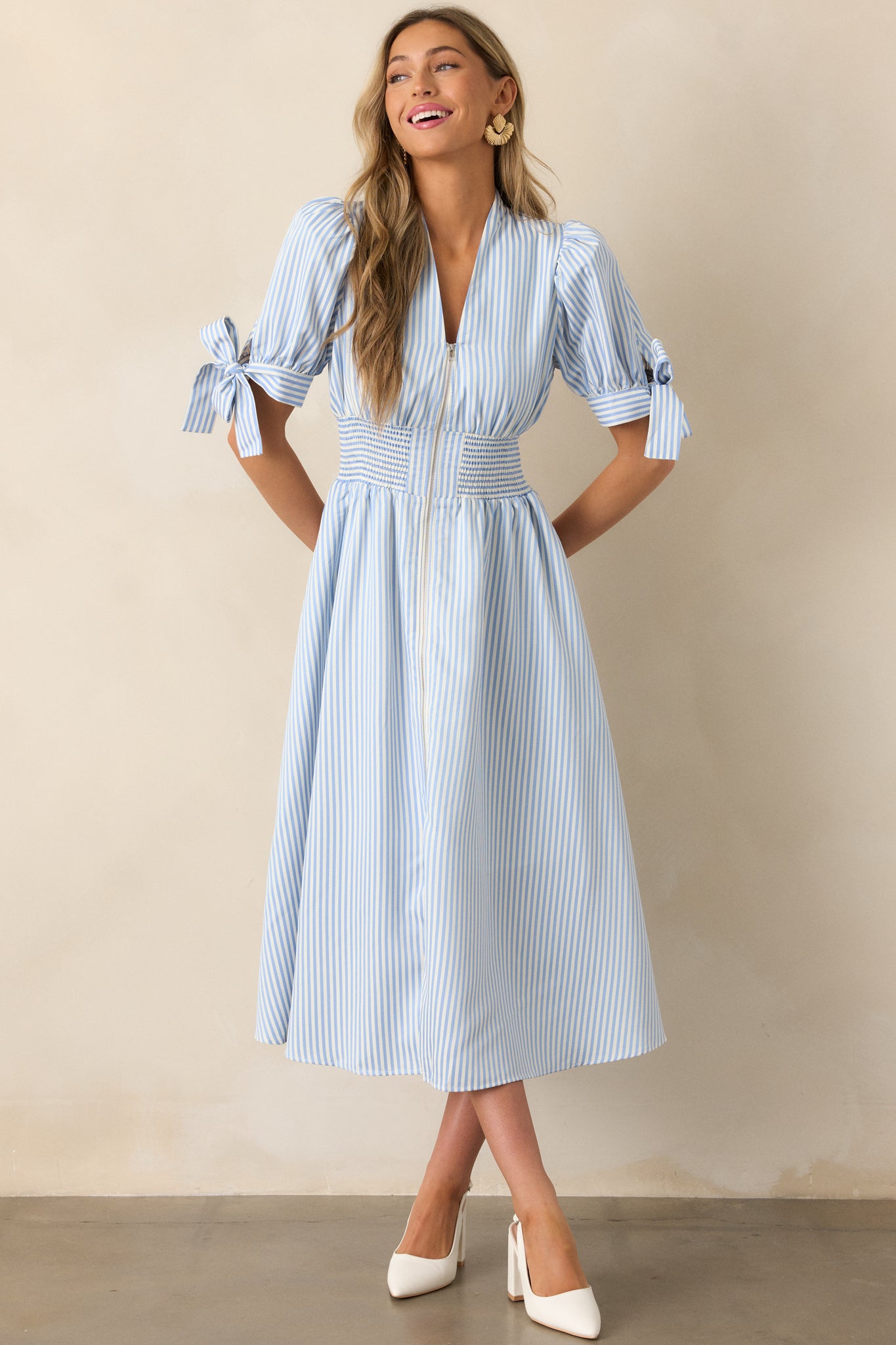 Blue stripe dress featuring a fitted waist with smocking detail, a V-neck neckline leading to a front zipper, and tie accents at the bottom of the sleeves.