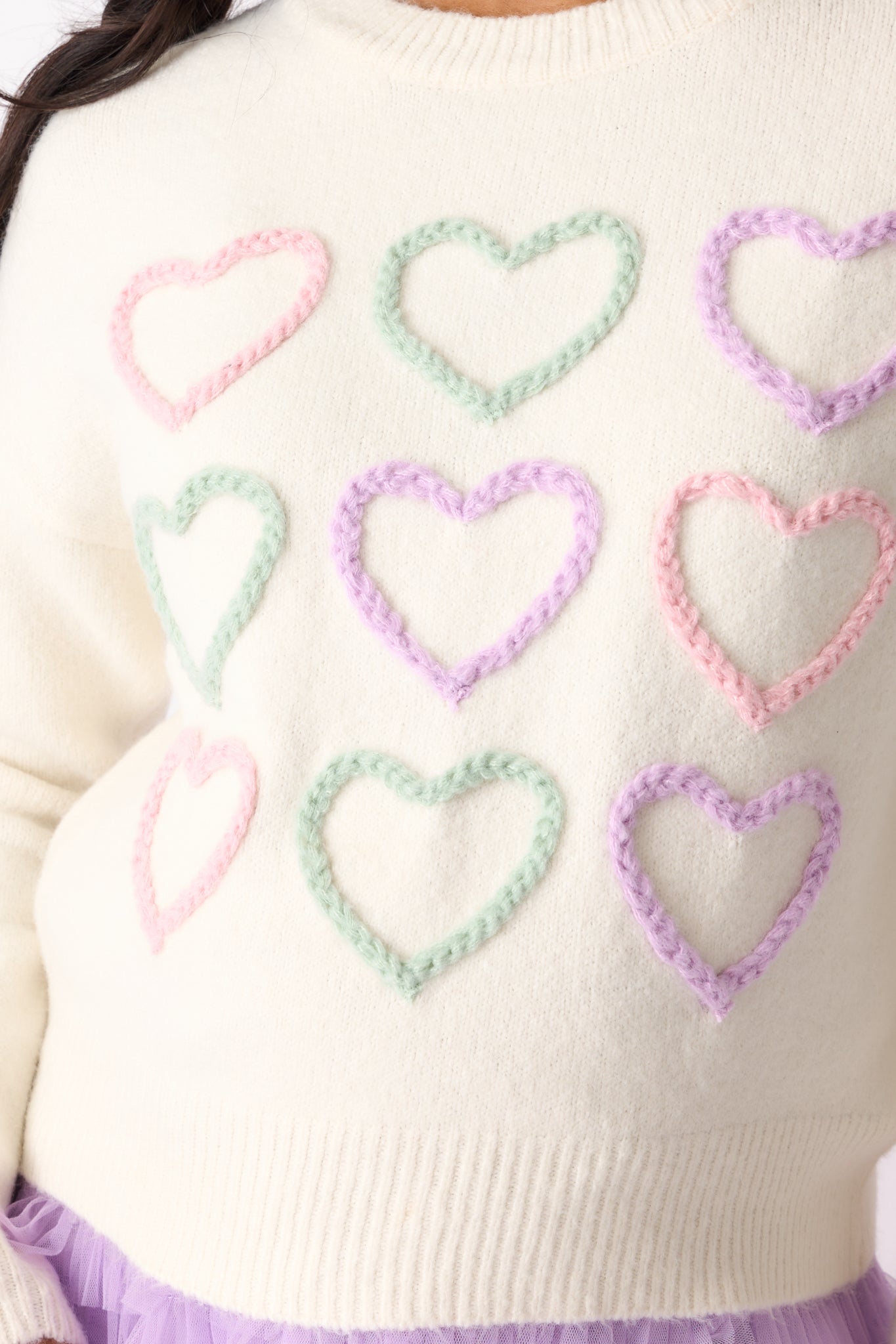 A detailed close-up showing the colorful heart stitch detail and smooth texture of the ivory sweater fabric.