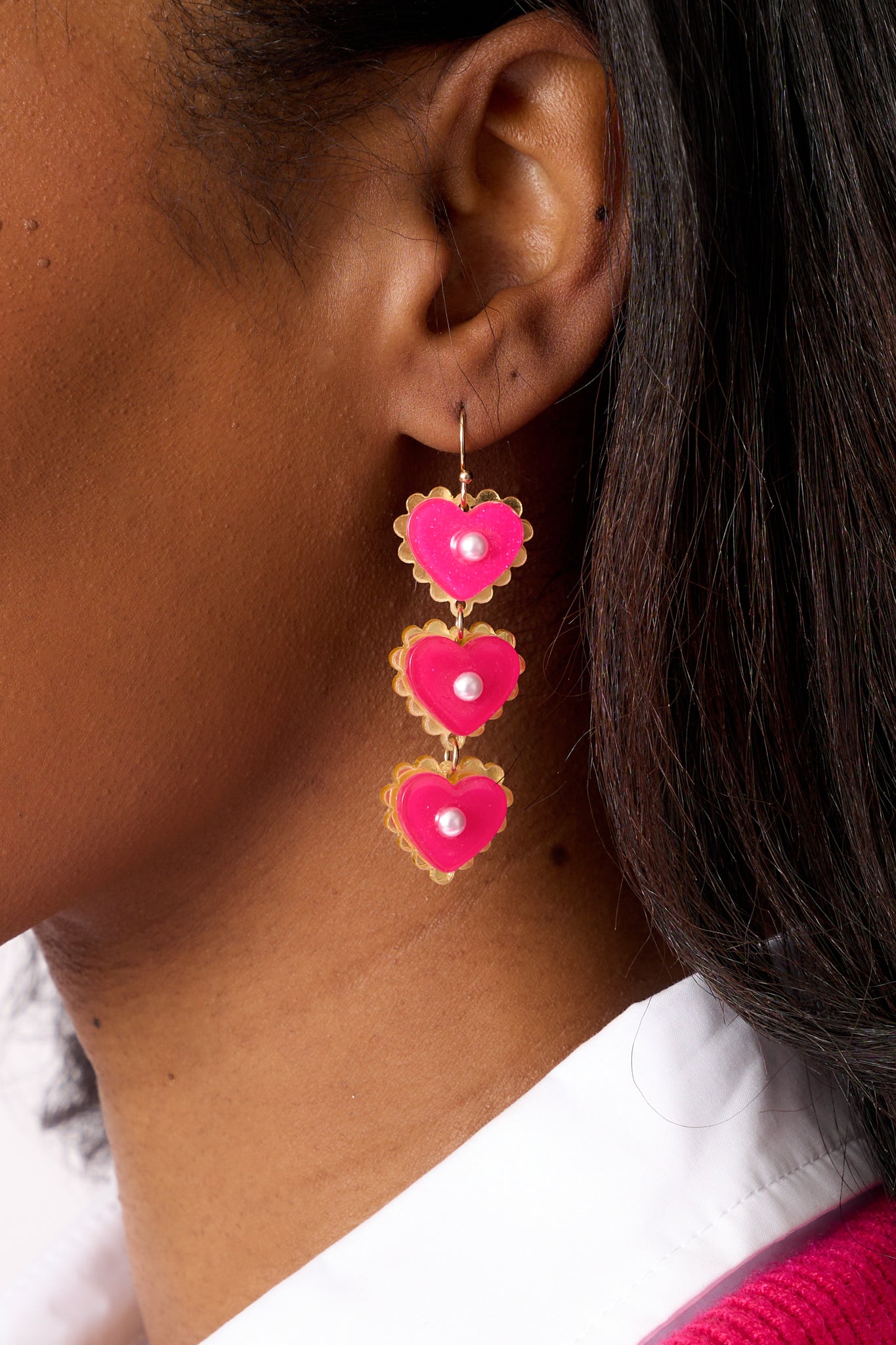 A side view of one pink heart drop earring, showing the three connected acrylic pink glitter heart charms with gold scallop detailing on the edges and a small ivory faux pearl centered in each heart.