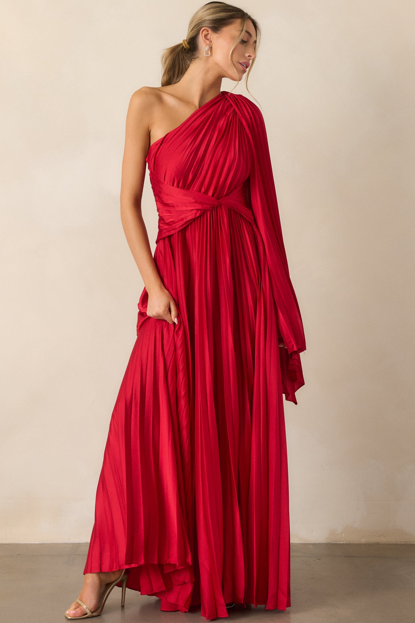 A full view of the red dress featuring a one-shoulder neckline, twist waist detailing, pleated fabric, and a flowing shoulder scarf accent.