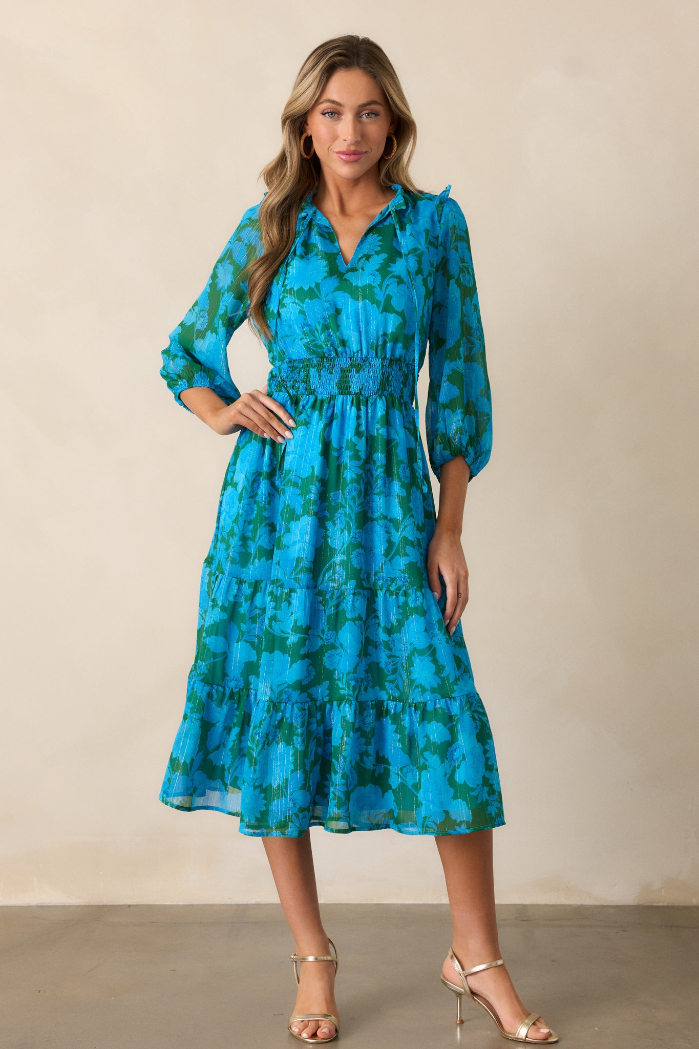 A full view of the blue dress showcasing its v-neckline with self-tie collar detailing, smocked waistband, and midi-length tiered hem skirt.