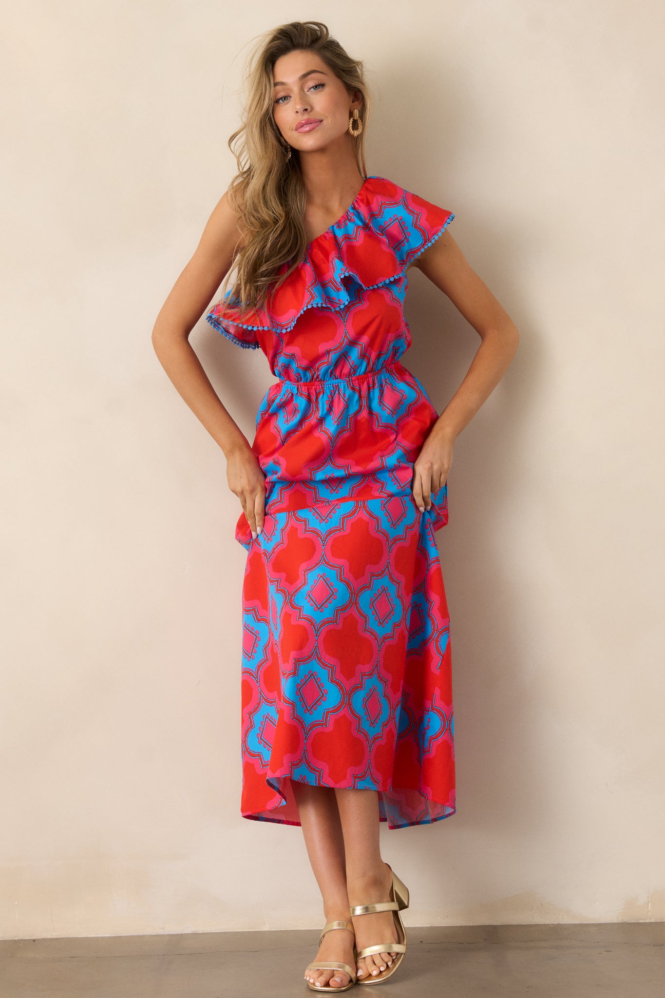  Full view of the red dress, featuring a one-shoulder design with a ruffle neckline accent and blue embroidered trim, paired with a midi length.