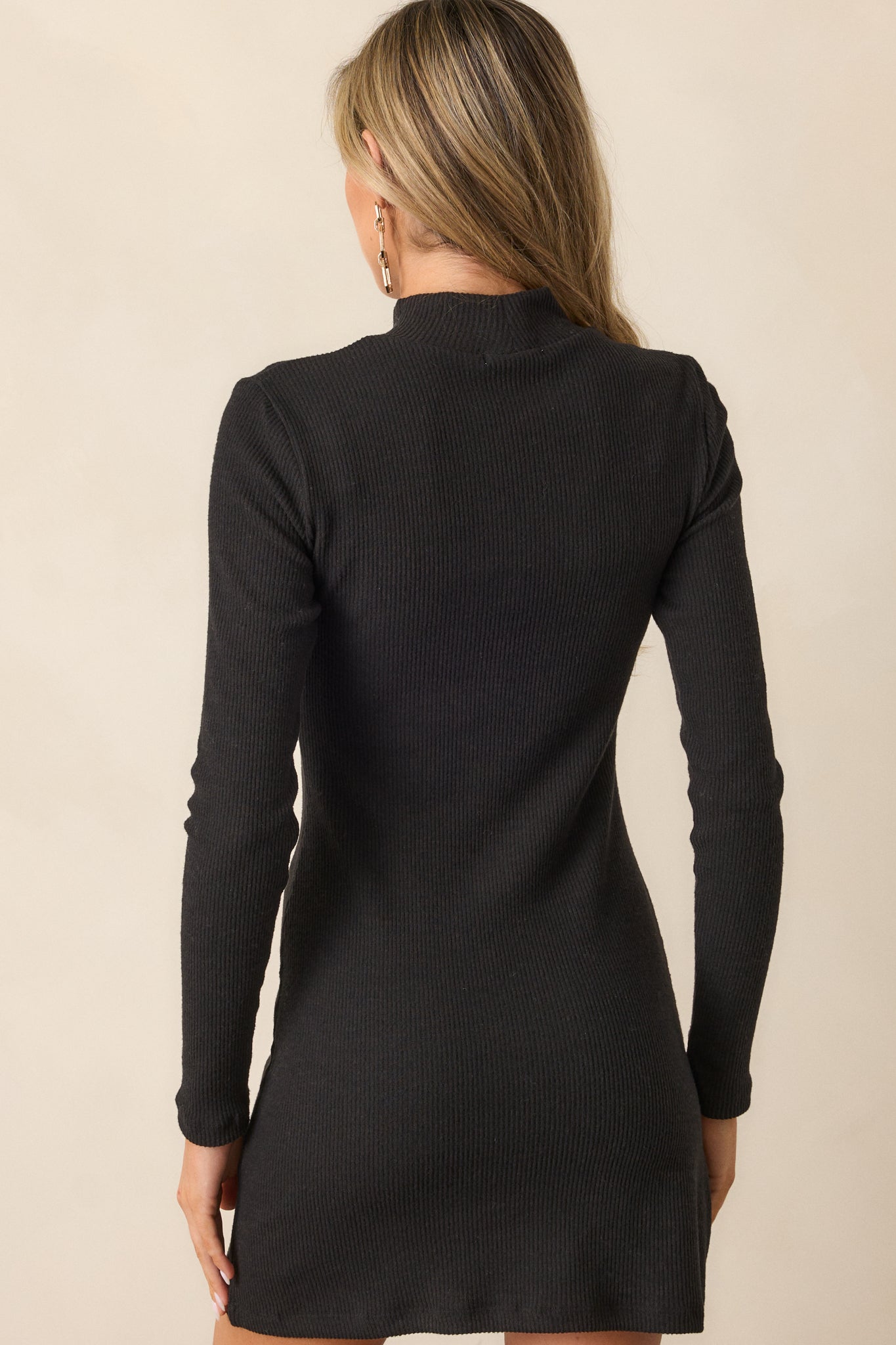 Back view of the black mini dress, highlighting the fitted design, ribbed fabric, and long sleeves with smooth detailing.