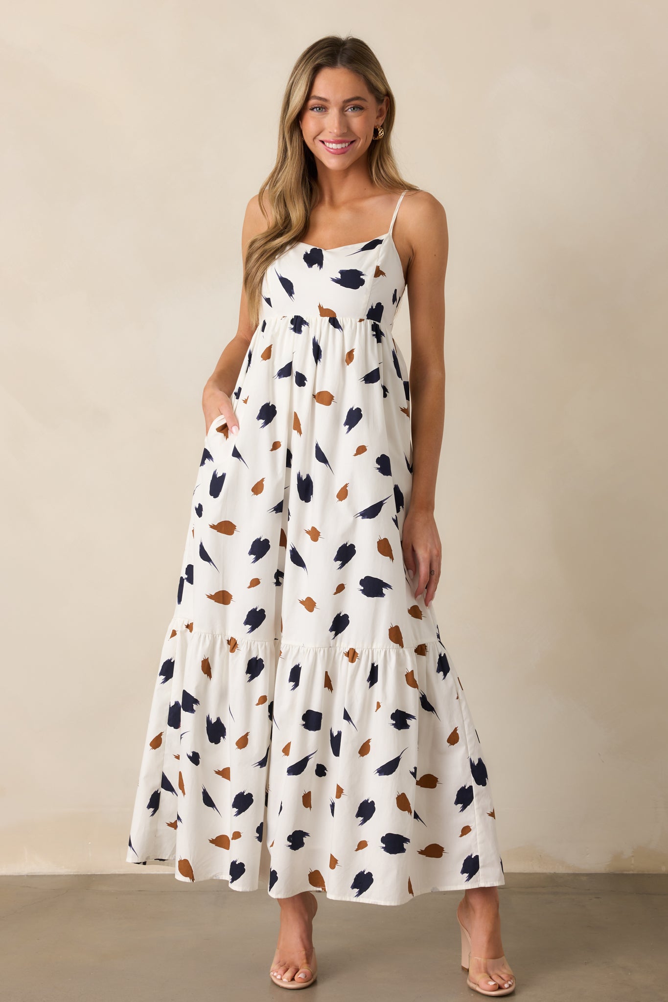 Ivory dress with a straight neckline, brown and navy brush stroke design, and a maxi length featuring adjustable spaghetti straps.