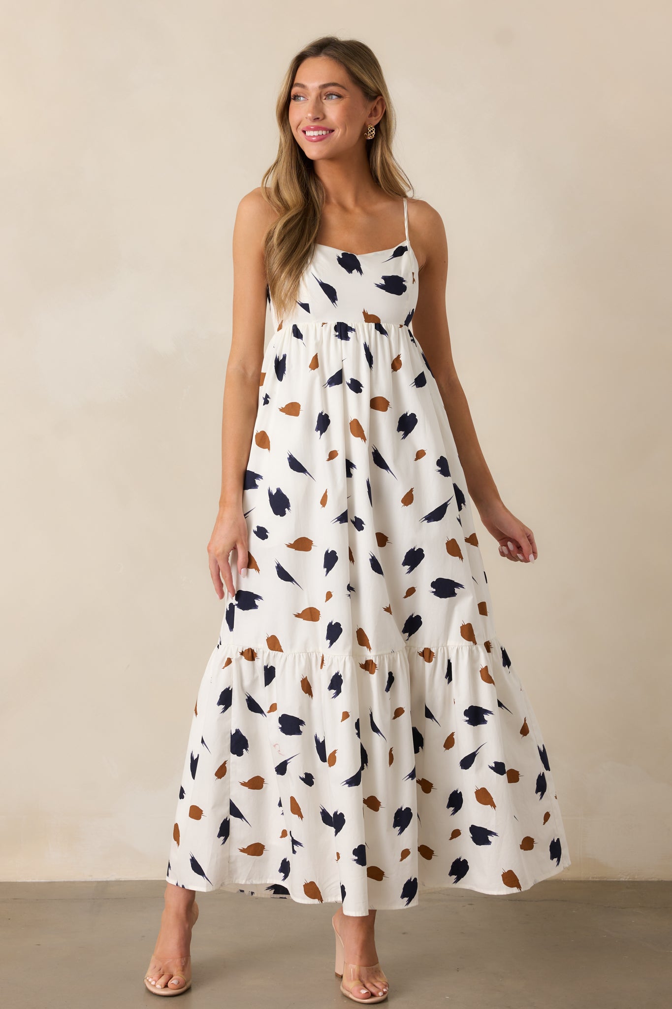 A flowing ivory maxi dress highlighted by a straight neckline, brown and navy brush stroke details, and an adjustable strap design.