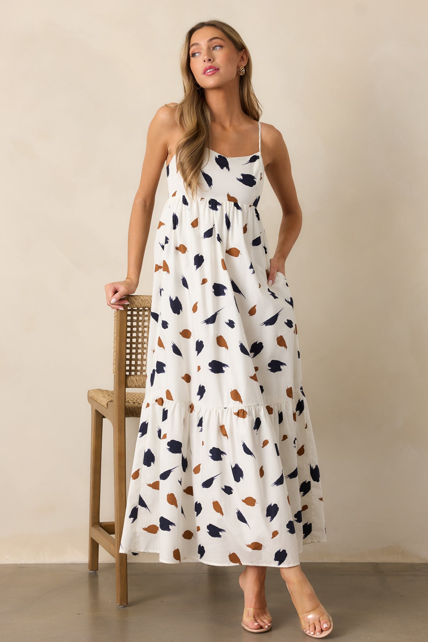 An ivory maxi dress with a straight neckline, adorned with a brown and navy brush stroke print, adjustable spaghetti straps, and a flowing silhouette.