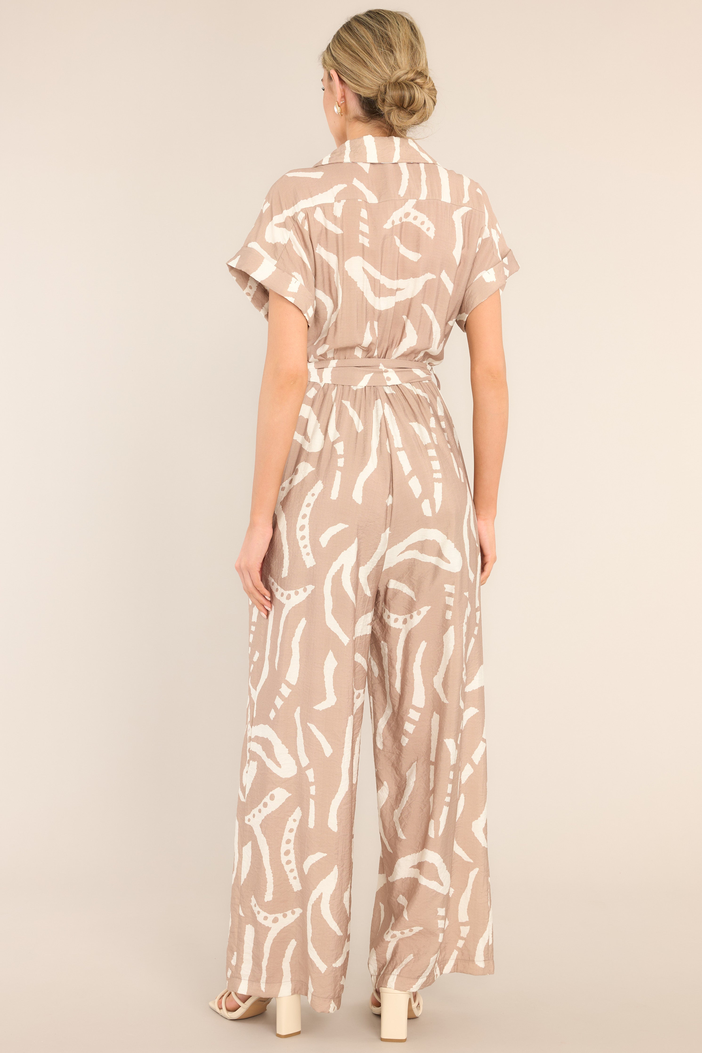 Back view of  this jumpsuit that features a notched lapel collared neckline, chest pockets, a functional button front, belt loops, a self-tie belt, an elastic insert at the back of the waist, side hip pockets, and a wide leg.