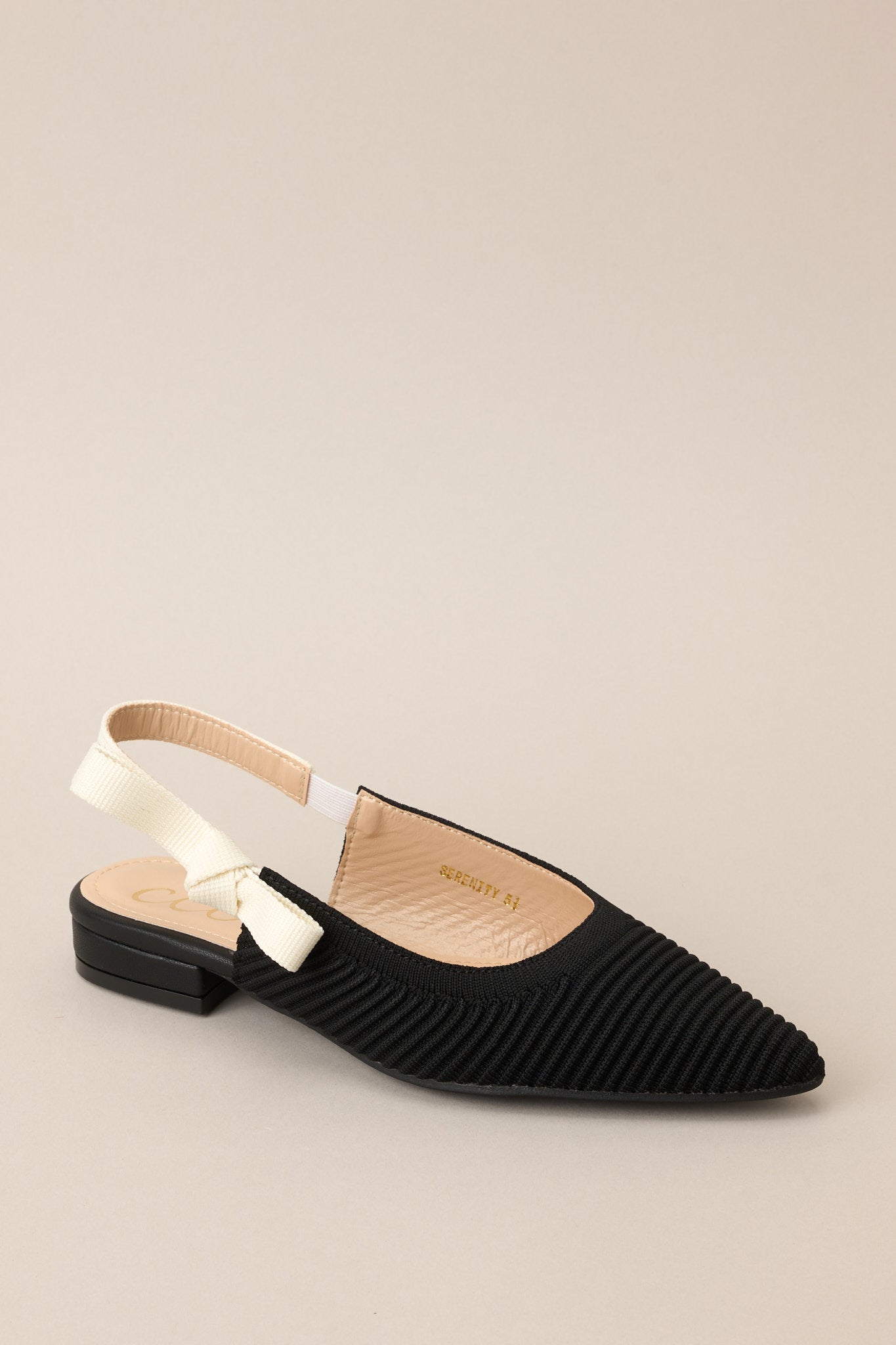 Angled side view of these shoes that feature a pointed toe, a textured material, a contrasting elastic strap around the back of the heel, and a subtle bow detail.