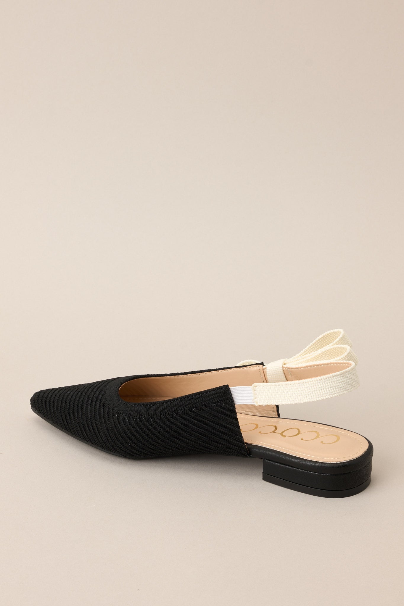 Inner-side view of these shoes that feature a pointed toe, a textured material, a contrasting elastic strap around the back of the heel, and a subtle bow detail.