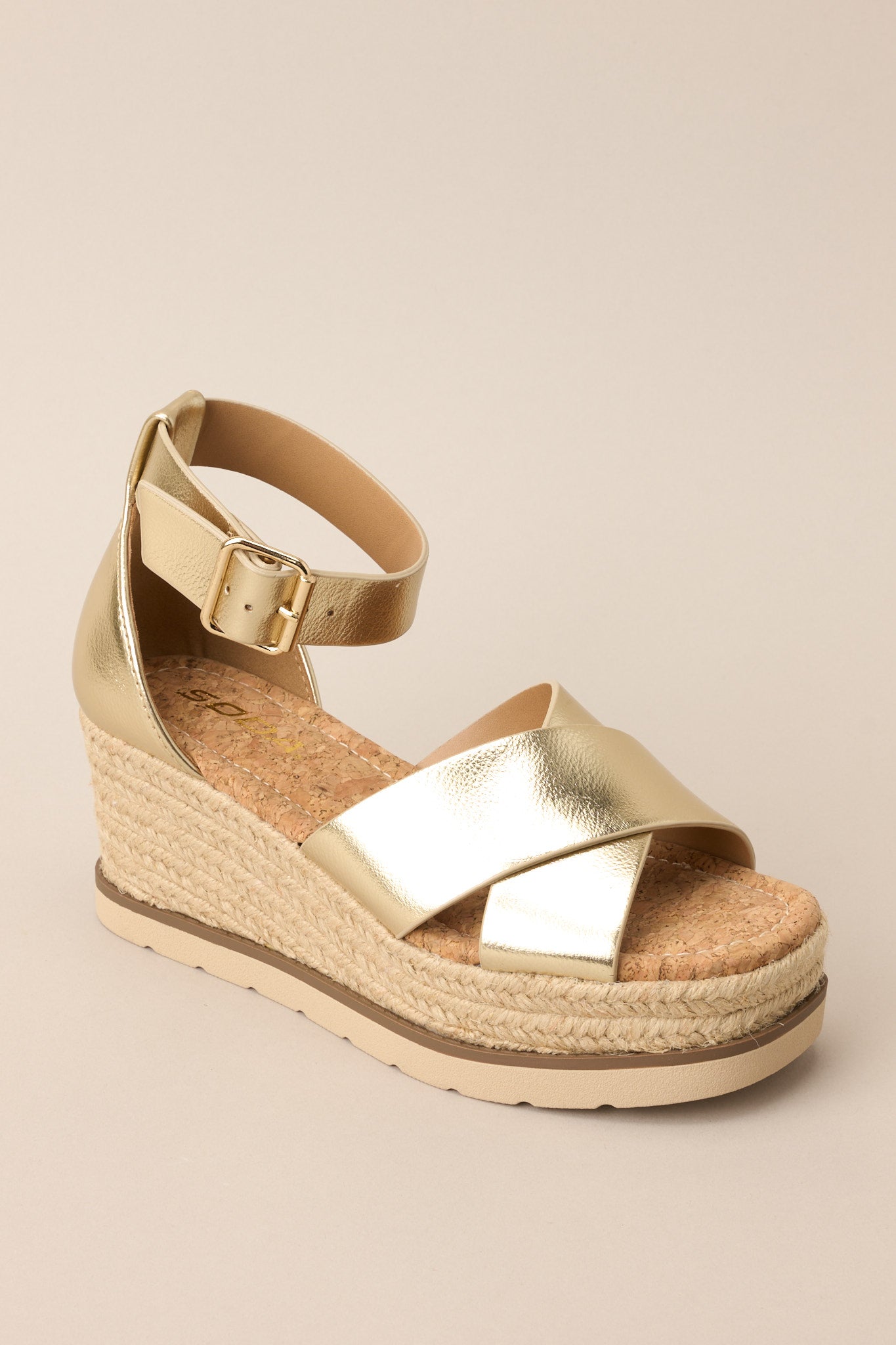 Angled front view of these sandals that feature a rounded toe, gold hardware, crossed straps over the top of the foot, an ankle strap with a classic buckle closure, and an espadrille wedged platform.