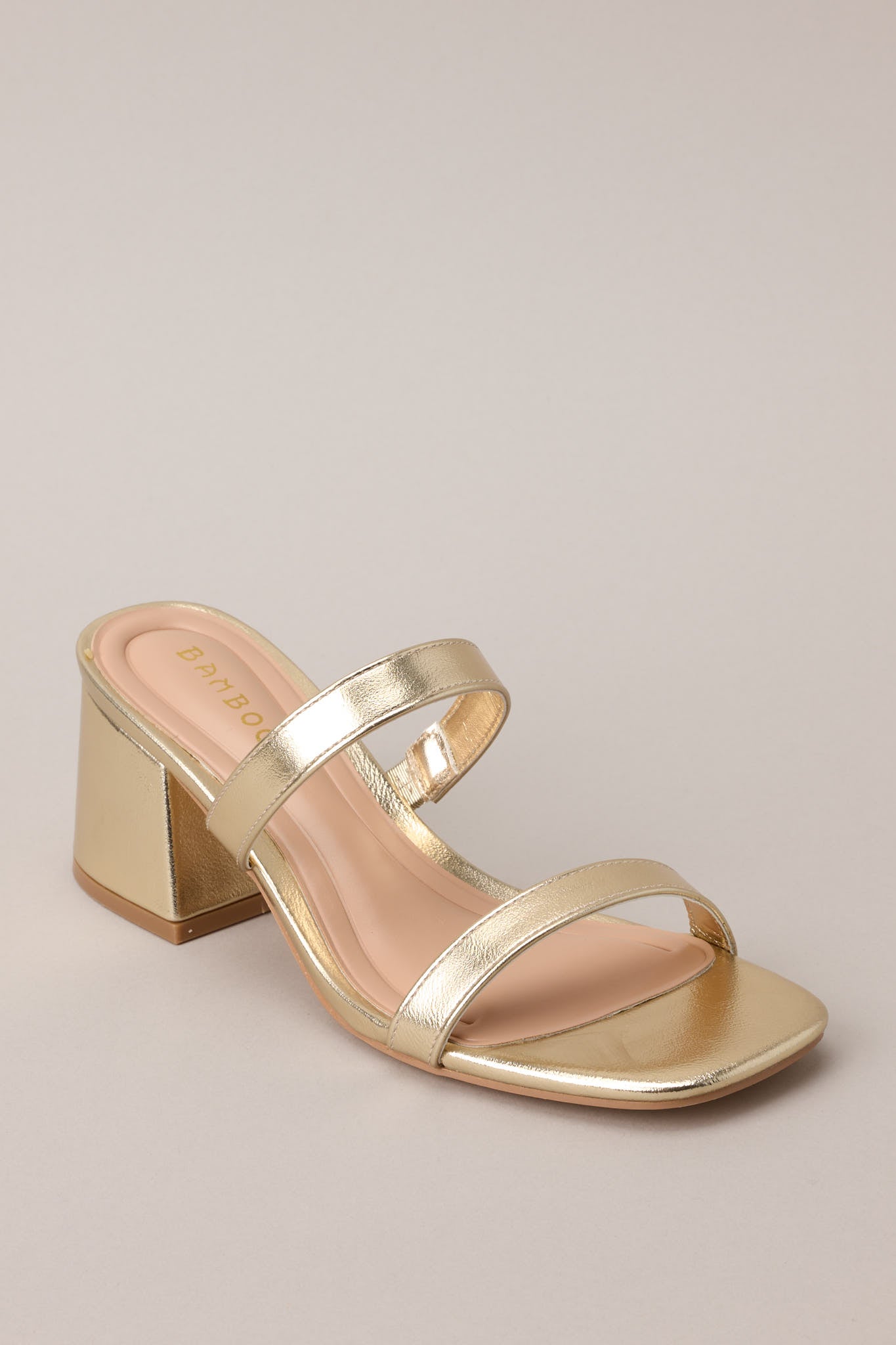 Angled close up view of these gold block heels that feature a square toe, two gold straps across the top of the foot, and a block heel.