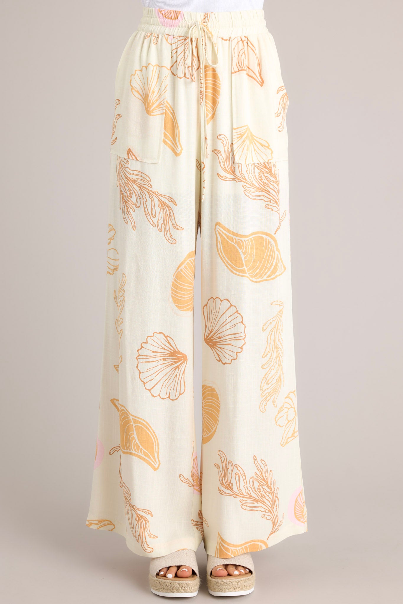 Full view of these ivory pants featuring an elastic waistband, a drawstring tie, a wide leg design, and a beachy seashell print throughout.