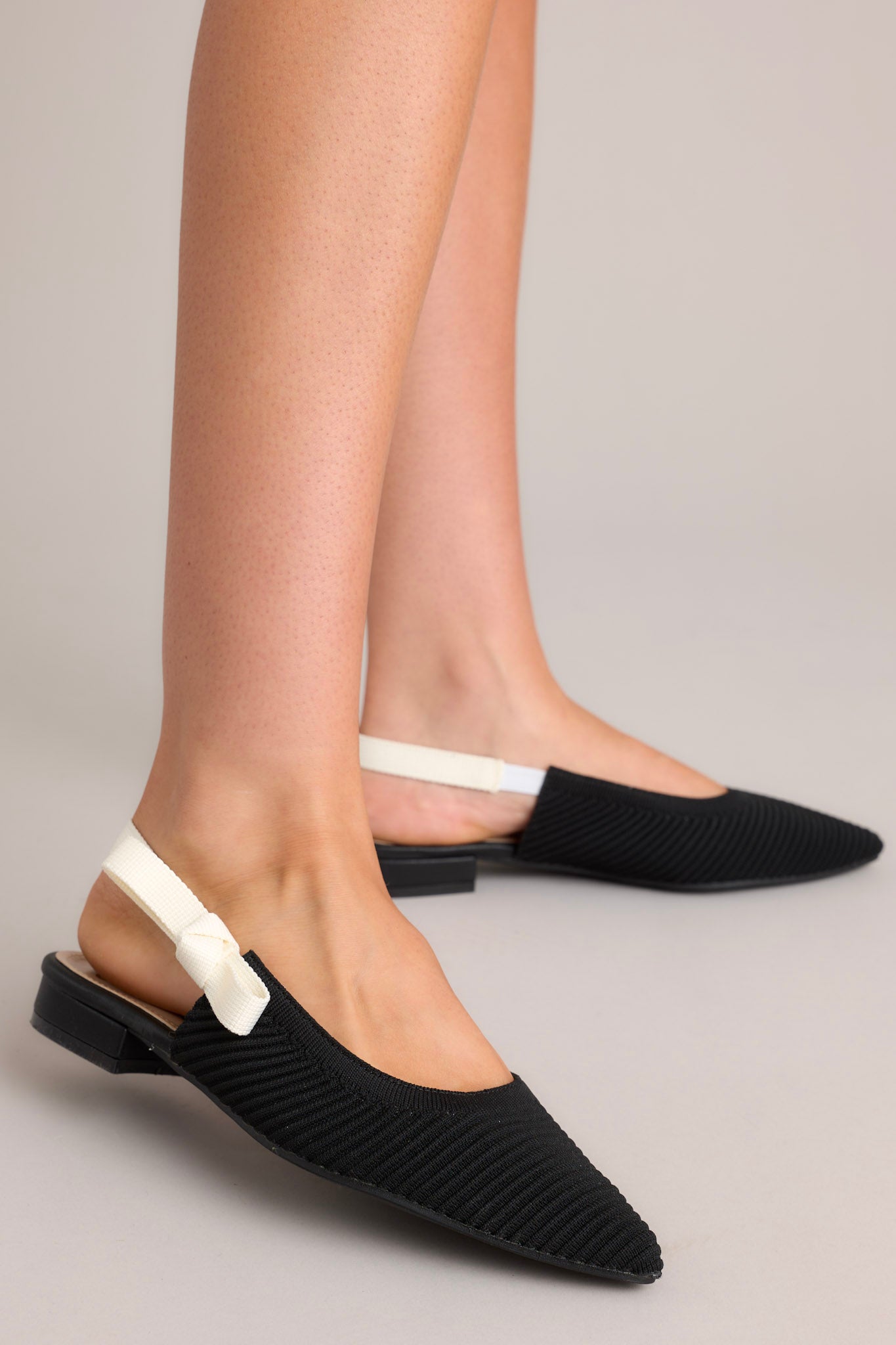 These black and white shoes feature a pointed toe, a textured material, a contrasting elastic strap around the back of the heel, and a subtle bow detail.