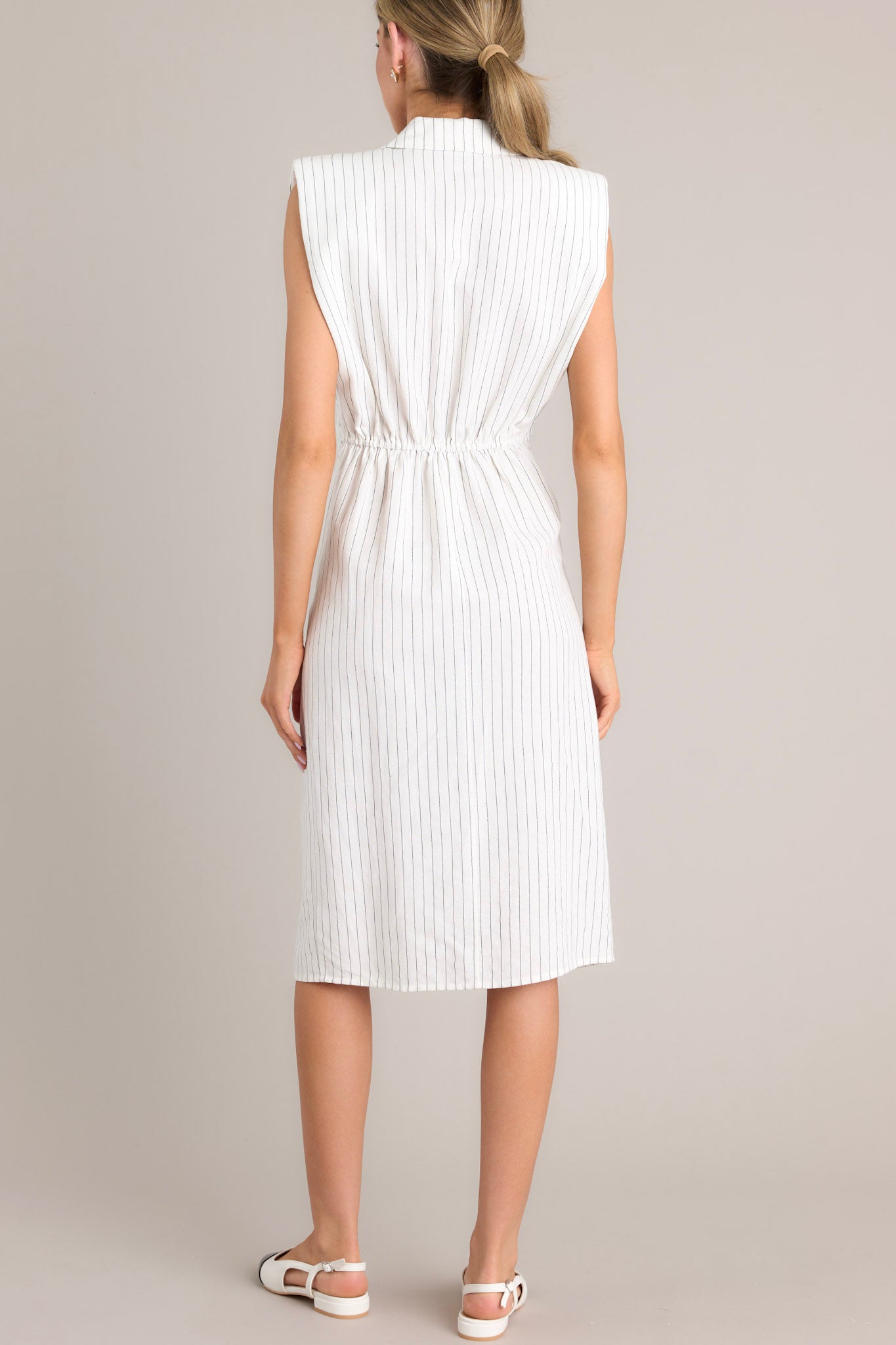 Back view of this pinstripe midi dress featuring a collared neckline, shoulder padding, a functional button front, a self-tie waist feature, an elastic insert at the back of the waist, and a front slit.