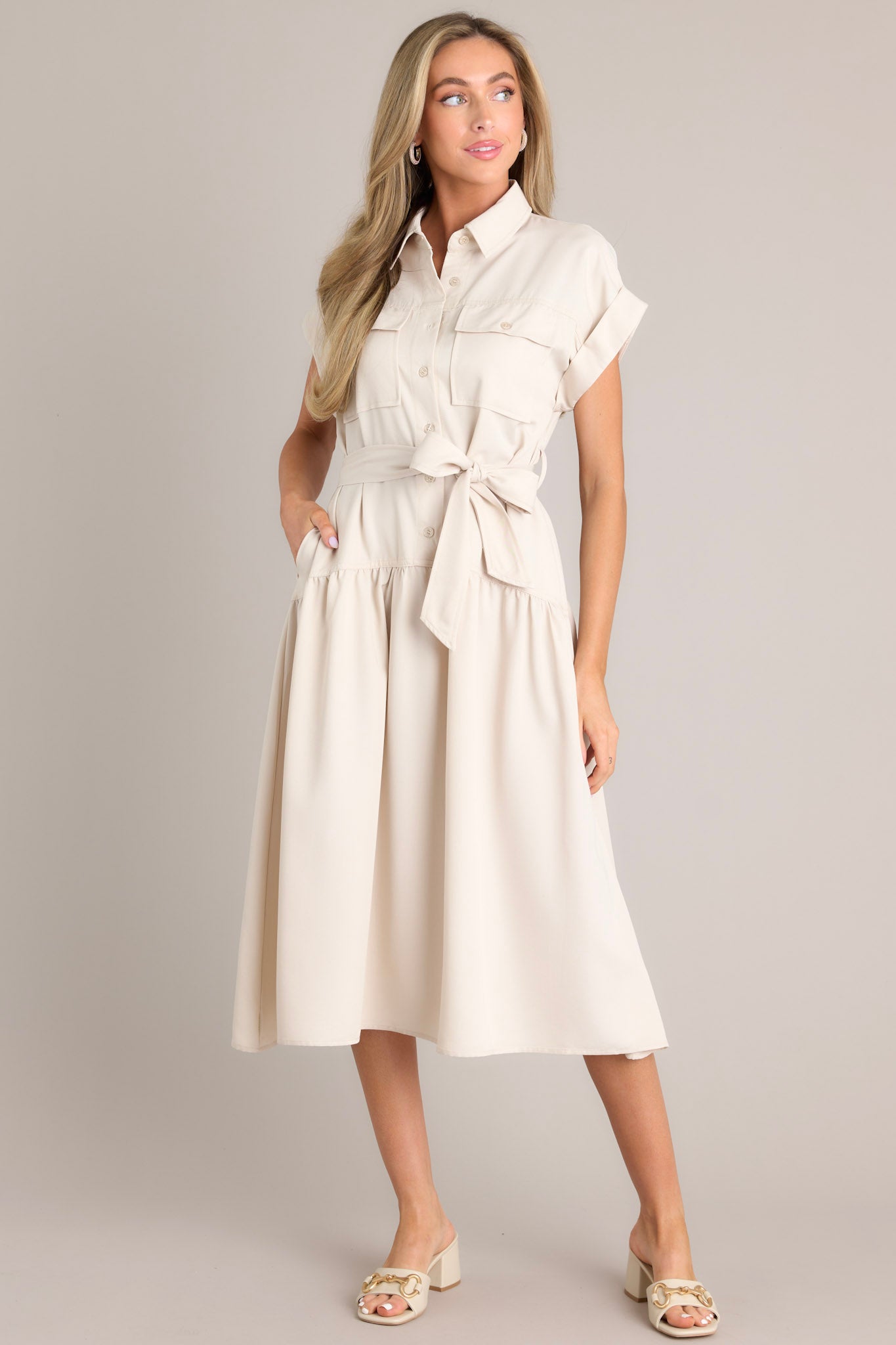 Angled view of this beige dress that features a collared neckline, functional breast pockets, a button front, a self-tie belt, belt loops, functional hip pockets, a single tier, and cuffed short sleeves.