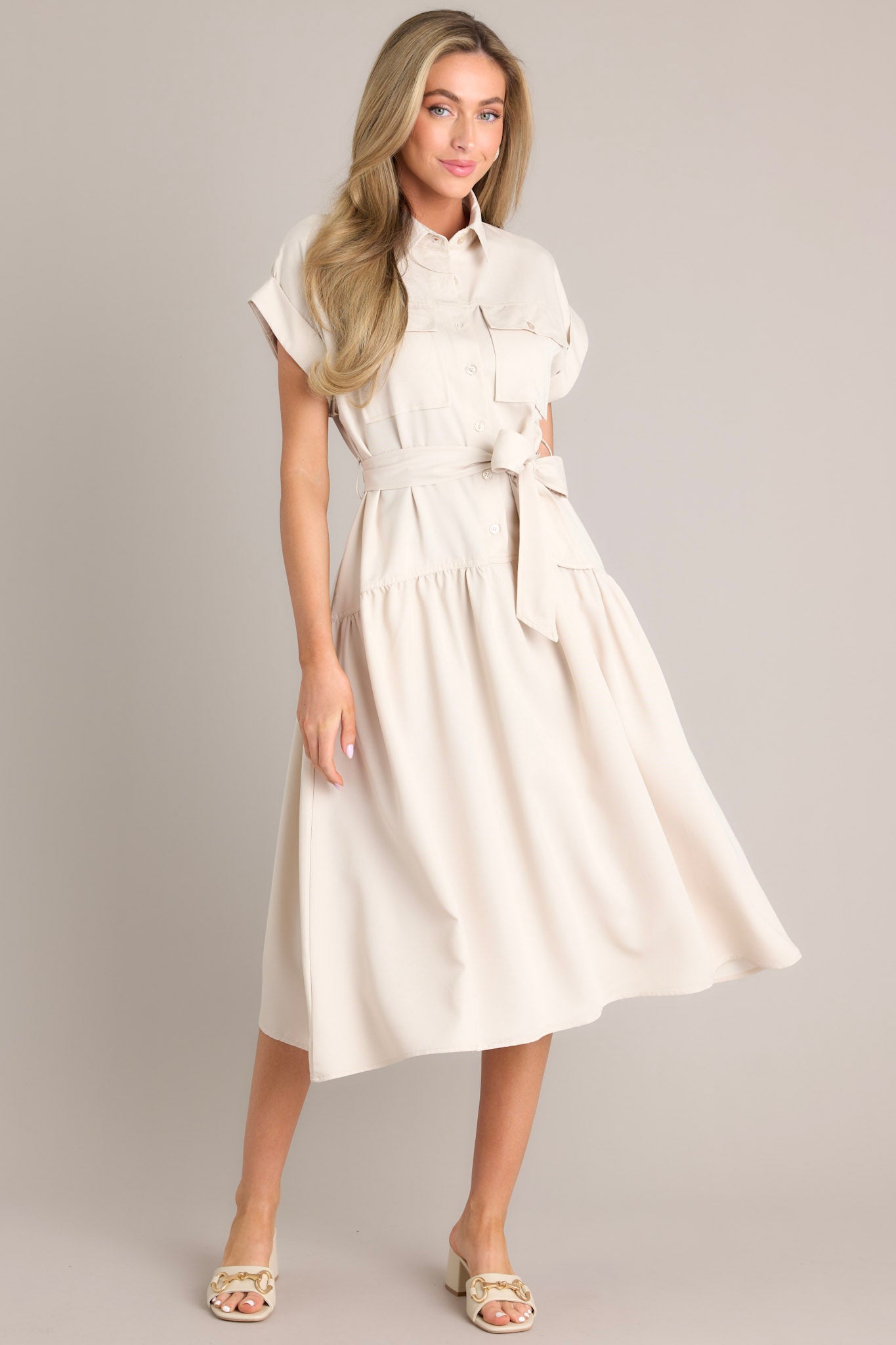 Full body view of this beige dress that features a collared neckline, functional breast pockets, a button front, a self-tie belt, belt loops, functional hip pockets, a single tier, and cuffed short sleeves.