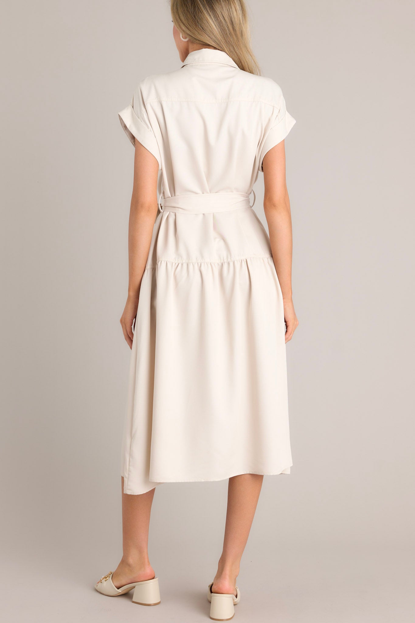 Back view of this beige dress that features a collared neckline, functional breast pockets, a button front, a self-tie belt, belt loops, functional hip pockets, a single tier, and cuffed short sleeves.