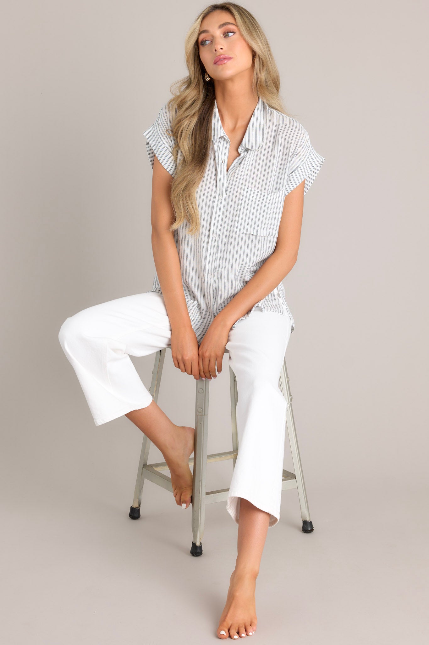 Seated full body view of this charcoal stripe top that features a collared neckline, a full button front, a functional breast pocket, and a split hemline.