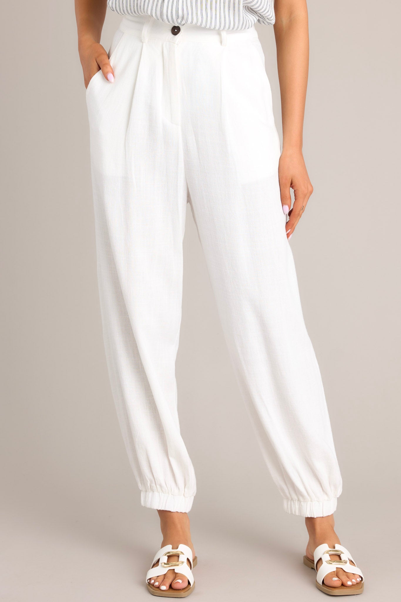 These white linen joggers feature a high rise design, a functional button & zipper closure, belt loops, subtle pleats, functional front & back pockets, and elastic cuffed ankles.