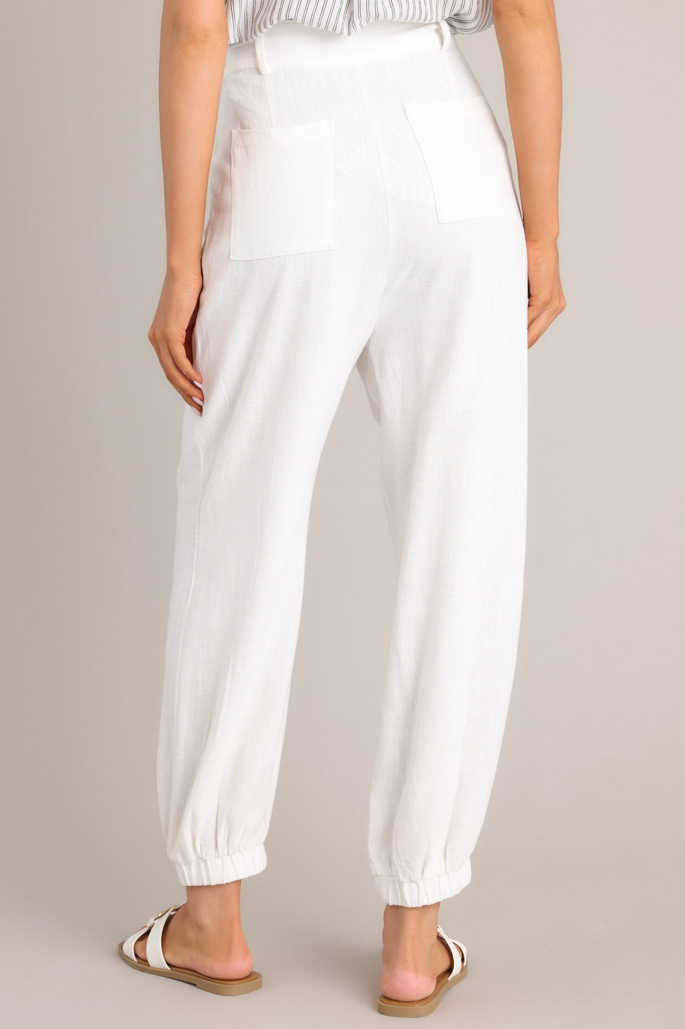 Back view of these white linen joggers that feature a high rise design, a functional button & zipper closure, belt loops, subtle pleats, functional front & back pockets, and elastic cuffed ankles.