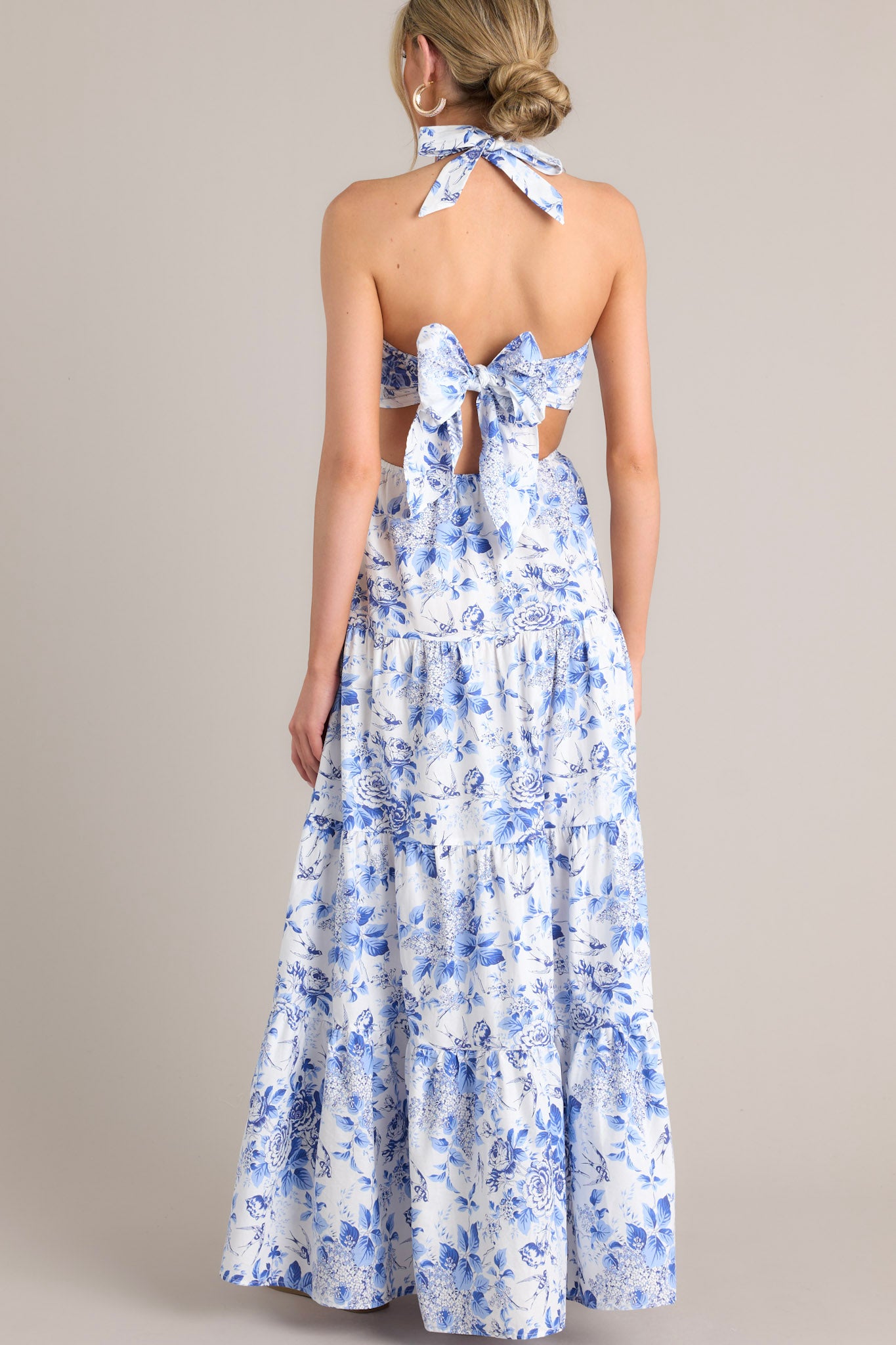 Full body back view of this floral maxi dress that features a self-tie halter neckline, an open back, a self-tie back feature, an elastic waistband, side cutouts, and a tiered design.