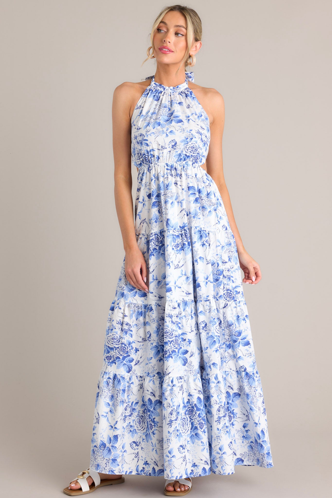 Angled view of this floral maxi dress that features a self-tie halter neckline, an open back, a self-tie back feature, an elastic waistband, side cutouts, and a tiered design.