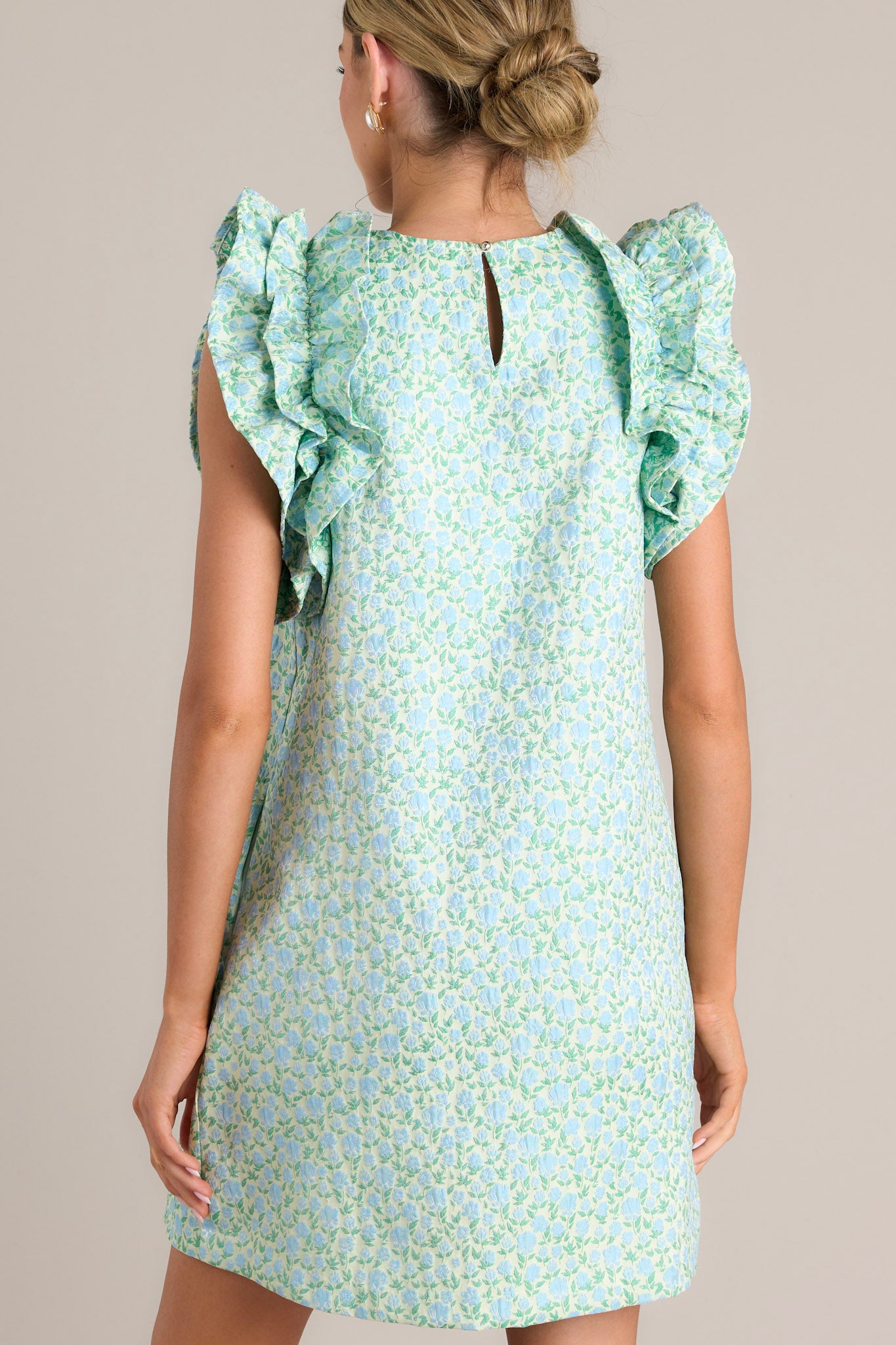 Back view of this light green mini dress that features a high crew neckline, a unique floral pattern, functional hip pockets, and ruffled sleeves.