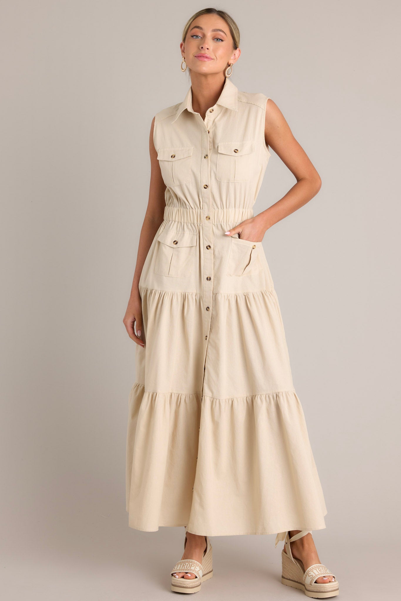 Angled full body view of this beige maxi dress that features a collared neckline, a full button front, buttoned breast pockets, a thick elastic waistband, buttoned hip pockets, and a tiered design.